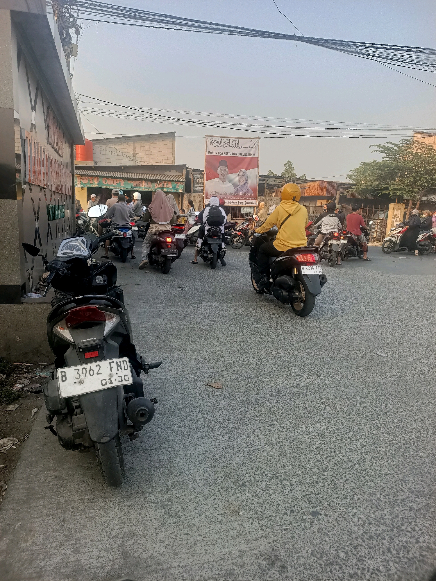 The image depicts a street scene in Wanajaya, Indonesia, characterized by a moderate amount of motorcycle traffic. In the foreground, a black scooter with a white license plate reading "B 3962 FND 01-30" is parked on the left side of the road, facing away from the viewer. Further down the street, several other motorcycles are visible, some with riders and some appearing to be moving in a procession or traffic. One rider in a bright yellow hoodie is prominently positioned on a motorcycle in the middle of the road, also facing away. The environment includes paved road surfaces, sidewalks with some debris, and buildings along the sides of the street. Overhead, a network of electrical wires is strung across the sky. A large banner with text and images of two individuals is displayed on a building in the background, suggesting a local event or advertisement. The lighting indicates it is either early morning or late afternoon, with a clear sky and soft sunlight.