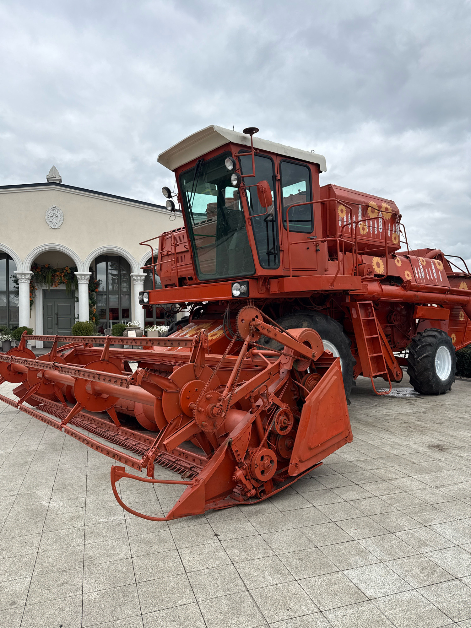 A red combine harvester is parked on a paved outdoor area in front of a building with arched windows and columns. The combine has a large harvesting head at the front and a cab with windows. Decorative sunflowers and wheat stalks are painted on the side of the combine. The sky is overcast. The scene is forensic, matter-of-fact, and devoid of any contextual information about location or specific purpose.