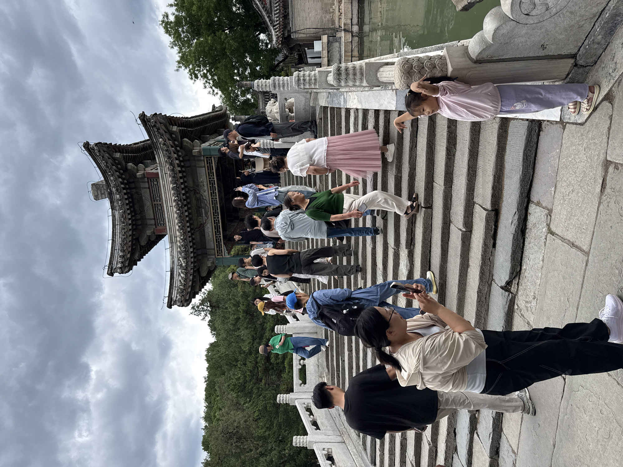 The image depicts a bustling outdoor scene at a multi-tiered traditional Chinese pavilion situated atop a wide stone bridge, likely within the Summer Palace (Yiheyuan) in Haidian District, China. A crowd of approximately 25-30 individuals, predominantly East Asian adults and several children, are ascending and descending the grey stone staircase that forms the bridge. The stairs feature ornate white stone balustrades with decorative pillars.
To the right of the bridge, a tranquil body of greenish water is visible, bordered by another stone structure. Dense green foliage and trees line the background on both sides, suggesting a natural park-like environment. The pavilion itself exhibits traditional Chinese architecture with multiple dark grey tiled roofs, upturned eaves, and visible wooden support structures. The sky above is heavily overcast with a uniform grey cloud cover.
In the immediate foreground, a woman with glasses and a ponytail, wearing a beige hoodie, light grey top, and black pants with white stitching, is holding a smartphone horizontally, appearing to capture media. To her right, a young girl in a light pink long-sleeved top and light purple pants leans against a bridge balustrade, striking a pose with both arms raised and bent at the elbows, looking towards the right side of the frame. Other individuals throughout the scene are walking, conversing, or observing their surroundings; several are holding smartphones, suggesting photography or communication. Clothing is casual, including jeans, t-shirts, hoodies, and comfortable footwear.
