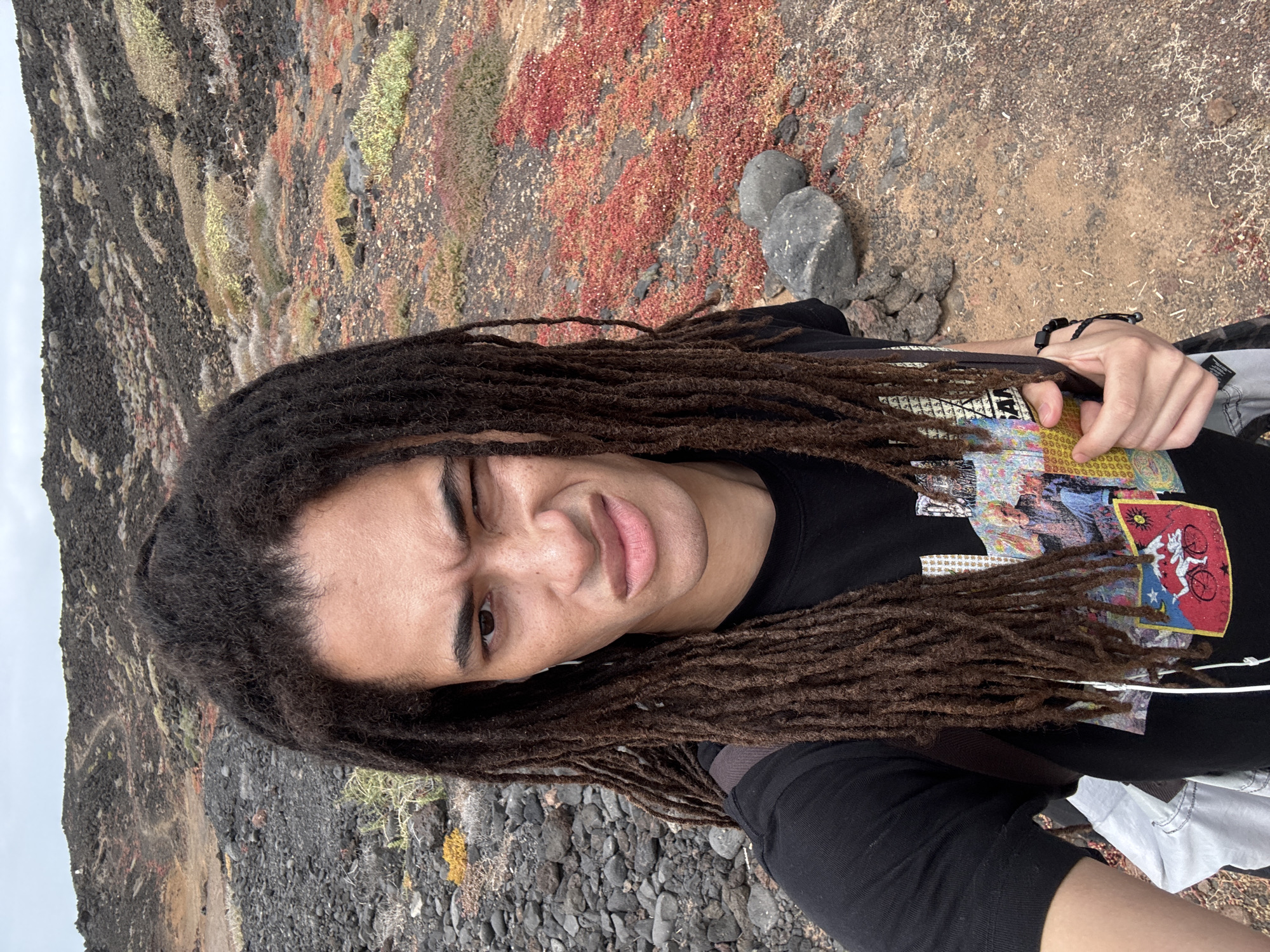 The image shows a person with dreadlocks in a selfie pose, wearing a black t-shirt with colorful graphic designs. The person's expression is slightly scrunched. The background reveals a volcanic landscape with red, brown, and black terrain, typical of the area around Tinajo, Spain. The ground is rocky with sparse vegetation.