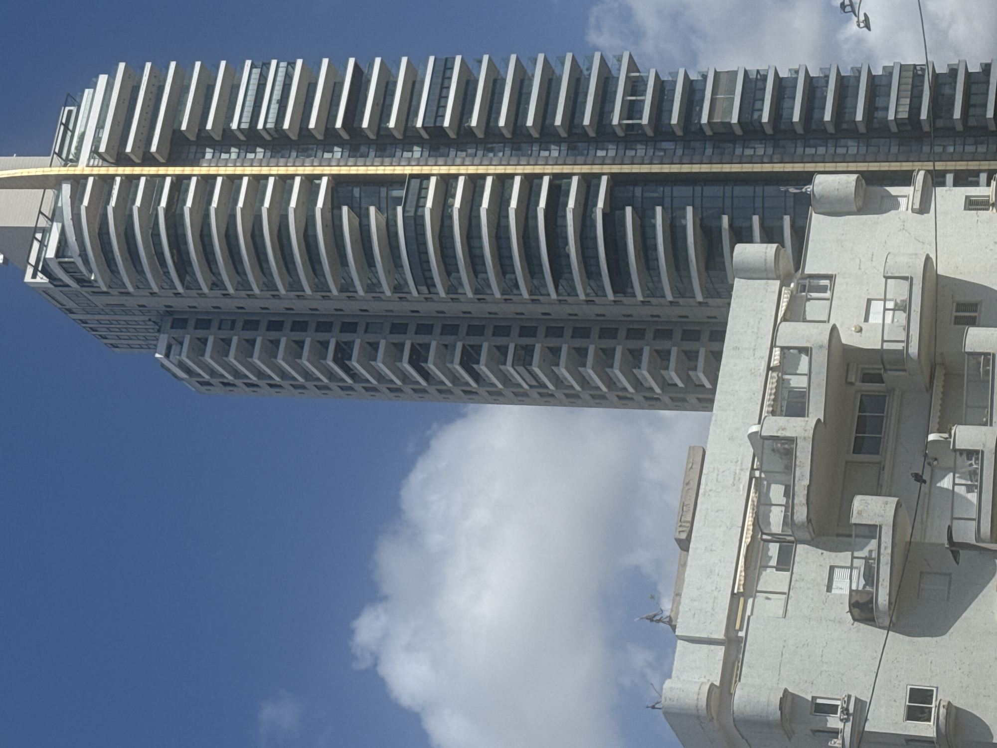 The image shows a low-angle view of two buildings against a blue sky with some white clouds. The building on the left is a tall, modern skyscraper with a facade of repeating geometric patterns, featuring vertical white slats and horizontal glazing. A gold-colored band runs horizontally through the middle of the building. To the right, there is an older, light-colored building with distinct architectural elements such as curved balconies and small rectangular windows. The city is Bat Yam, Israel.