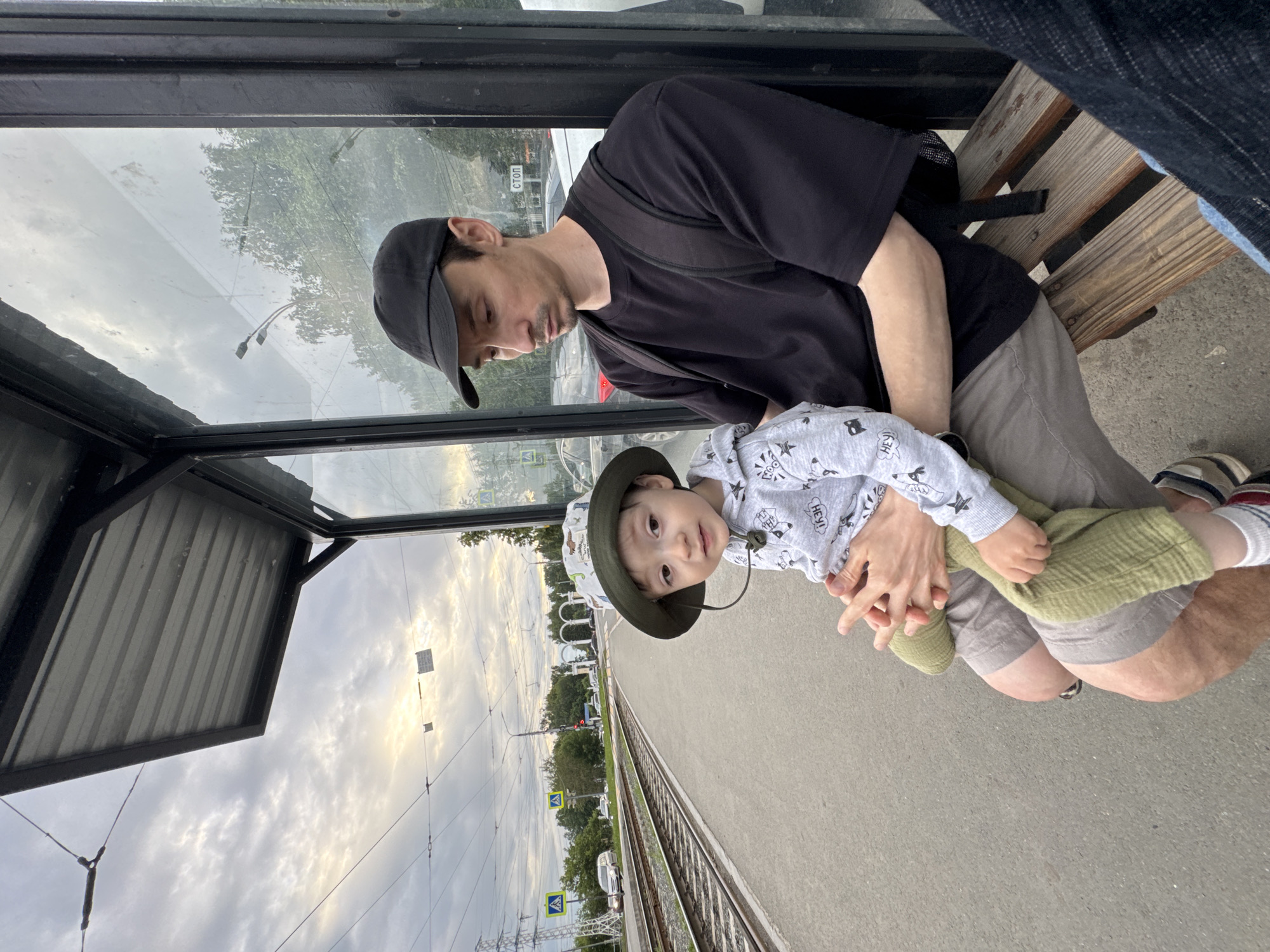 At a tram stop in Izhevsk, Russia, a man and a young child sit on a wooden bench under a metal-roofed shelter with a glass wall. The man, wearing a black t-shirt and a black baseball cap, holds the child on his lap. The child, wearing a patterned bucket hat, a grey hooded sweatshirt, and green pants, looks towards the camera. In the background, tram tracks run alongside a concrete platform. Overhead, power lines crisscross the sky, which is filled with clouds. Beyond the tracks, trees and buildings are visible, along with streetlights and traffic signals. A sign with Russian text, "СТОП" (STOP), is mounted on a pole. The scene appears to be set in the late afternoon or early evening, given the lighting conditions.