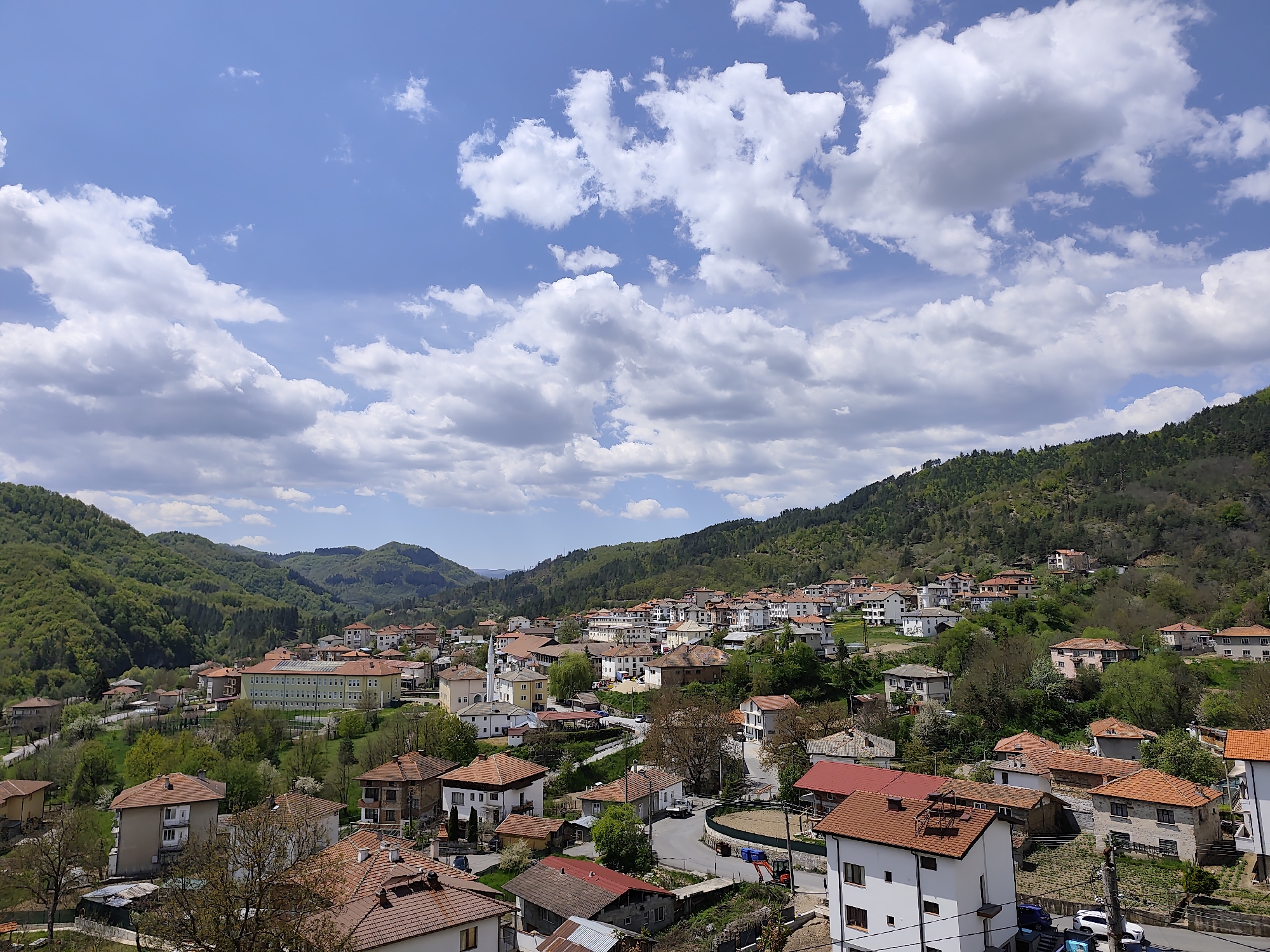 The image depicts the city of Srednogortsi, Bulgaria, on a sunny day with a partially cloudy sky. The town is nestled among green, rolling hills, with a mix of deciduous and coniferous trees covering the slopes. Buildings are predominantly white or light-colored with orange-red tiled roofs. In the center is a large, pale yellow building with multiple floors and evenly spaced windows. Other buildings are scattered across the landscape, creating a village-like atmosphere. In the immediate foreground, are structures with visible electrical wires running nearby. The sky features blue patches interspersed with cumulus clouds, creating a dynamic atmosphere above the town. The overall impression is a tranquil and picturesque mountain town in Bulgaria.