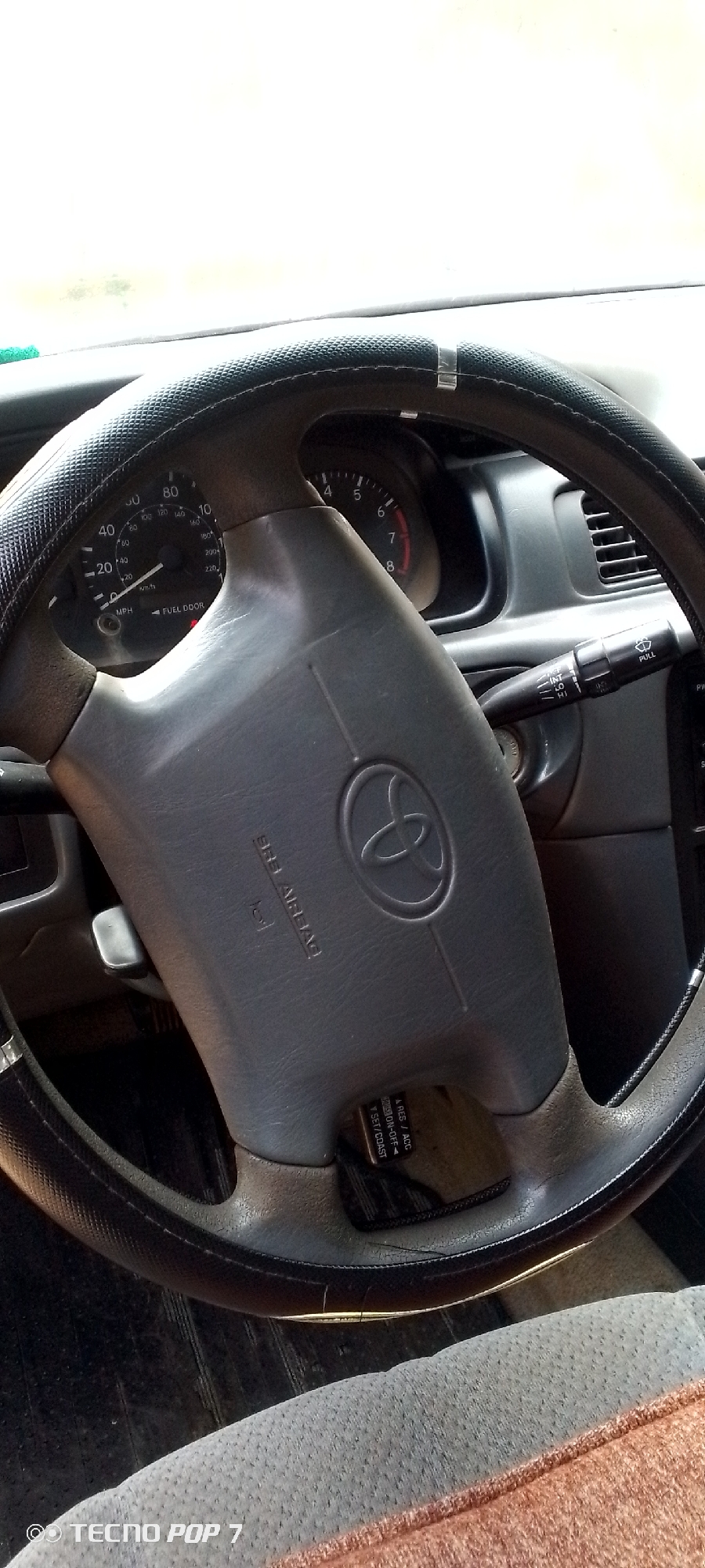 The image shows a close-up view of a car's interior, focusing on the steering wheel and dashboard. The steering wheel is black with a textured grip and has a Toyota logo in the center, along with "SRS AIRBAG" text. To the right of the steering wheel, a stalk with controls for windshield wipers is visible, as are the car's instrument cluster and part of the center console. The instrument cluster shows a speedometer with readings in MPH and KM/H, and a fuel gauge. A portion of the passenger seat, upholstered in a textured fabric, is visible in the foreground. The environment appears to be indoors or in a shaded area, given the lighting. No people are visible in the image.