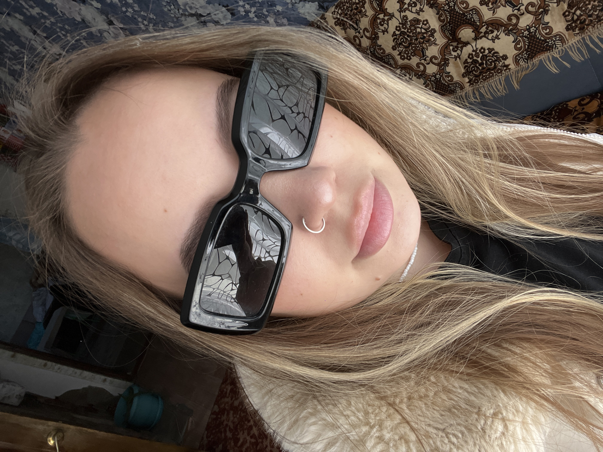 The image depicts a fair-skinned young woman with blonde hair wearing black sunglasses and a silver nose ring. She is lying down, with her head tilted towards the right. The background includes a patterned throw blanket and a glimpse of a room with a wooden dresser and a small blue object. No evidence in the picture reveals the location in the city of Novopokrovskaya, Russia.