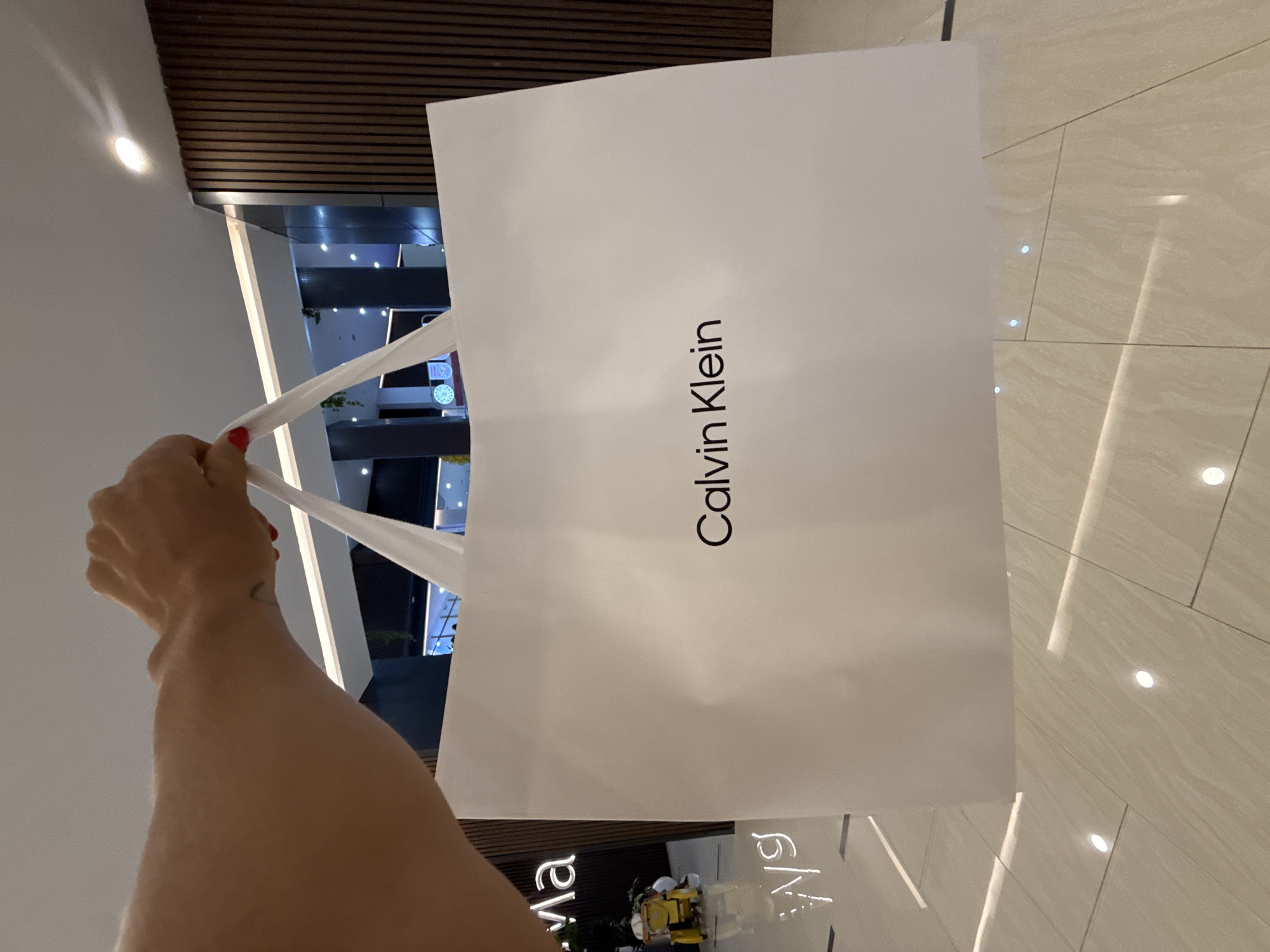 A person's arm and hand are holding a white Calvin Klein shopping bag with black lettering. The background shows elements of a modern indoor shopping mall. The location is Kuta, Indonesia as seen through the reflective floor spelling "Beachwalk".