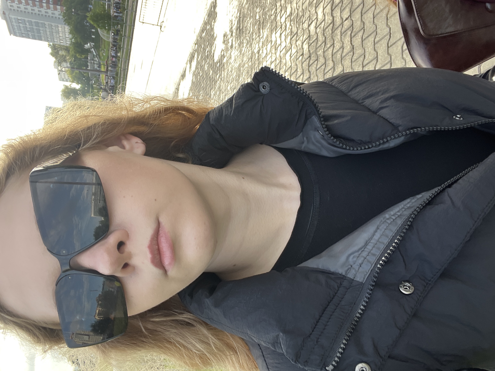 A young woman with blonde hair stands outdoors in a city, possibly Perm, Russia. She is wearing large, dark sunglasses that reflect the sky and buildings. She has on a black puffer jacket zipped over a black shirt, and a small hoop earring is visible in her left ear. The background shows buildings, trees, and a street or walkway paved with interlocking bricks, suggesting an urban park or public space. The lighting indicates it might be late afternoon or early morning, with long shadows cast by the sun.