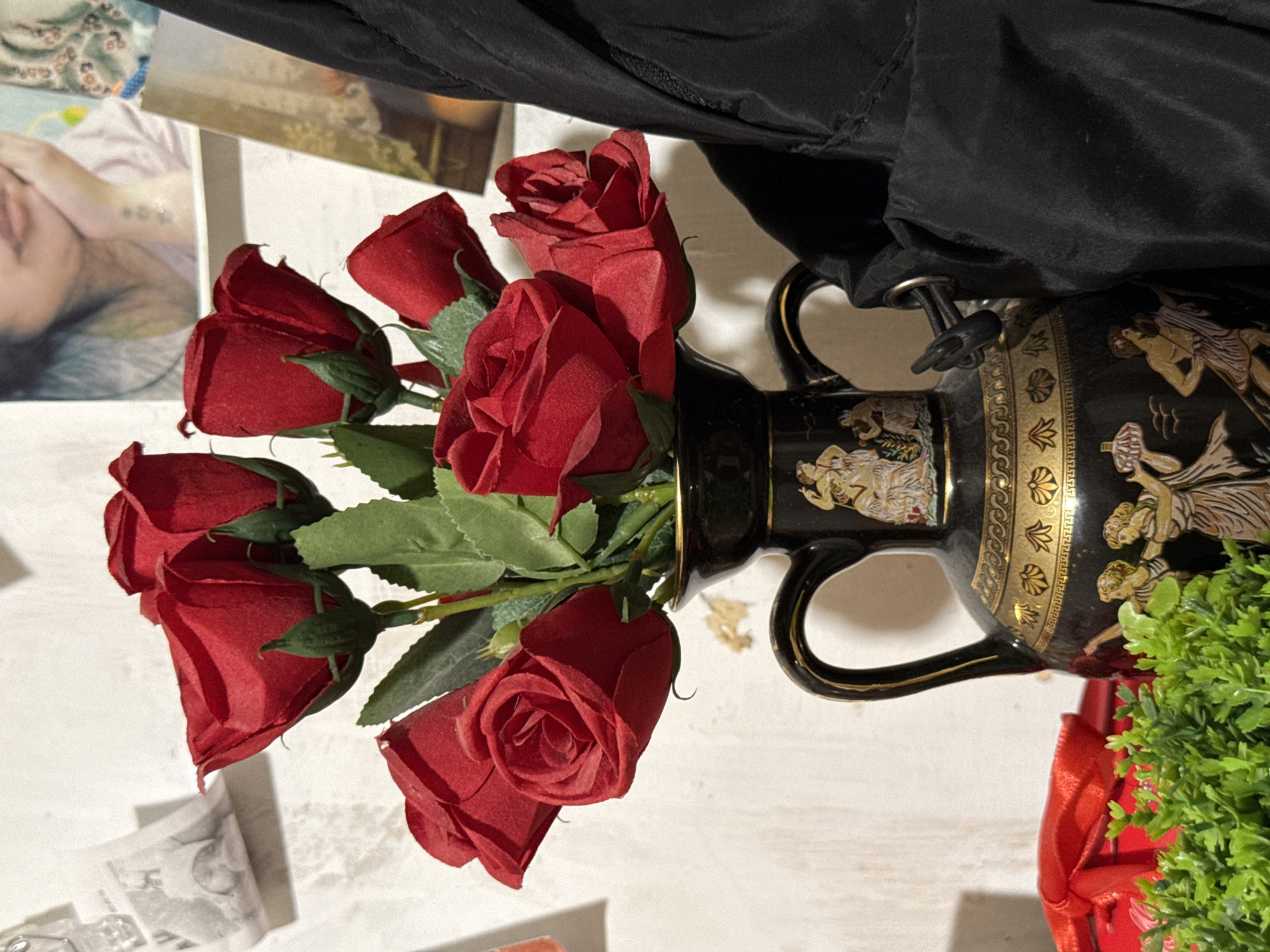 A bouquet of seven artificial red roses is arranged in a decorative black vase with gold trim. The vase features painted figures in classical style. The roses are positioned towards the left side of the frame, with some leaves visible. To the left of the vase, a red gift box with a ribbon is partially visible, and in front of the vase is a cluster of green artificial plants. In the background, against a textured, light-colored wall, are several framed pictures or prints. A black garment, possibly a jacket or bag, is positioned to the right of the vase, partially obscuring the background. The scene appears to be indoors, in a domestic setting. There is no indication of the city of Nyurba, Russia, or any specific location details.