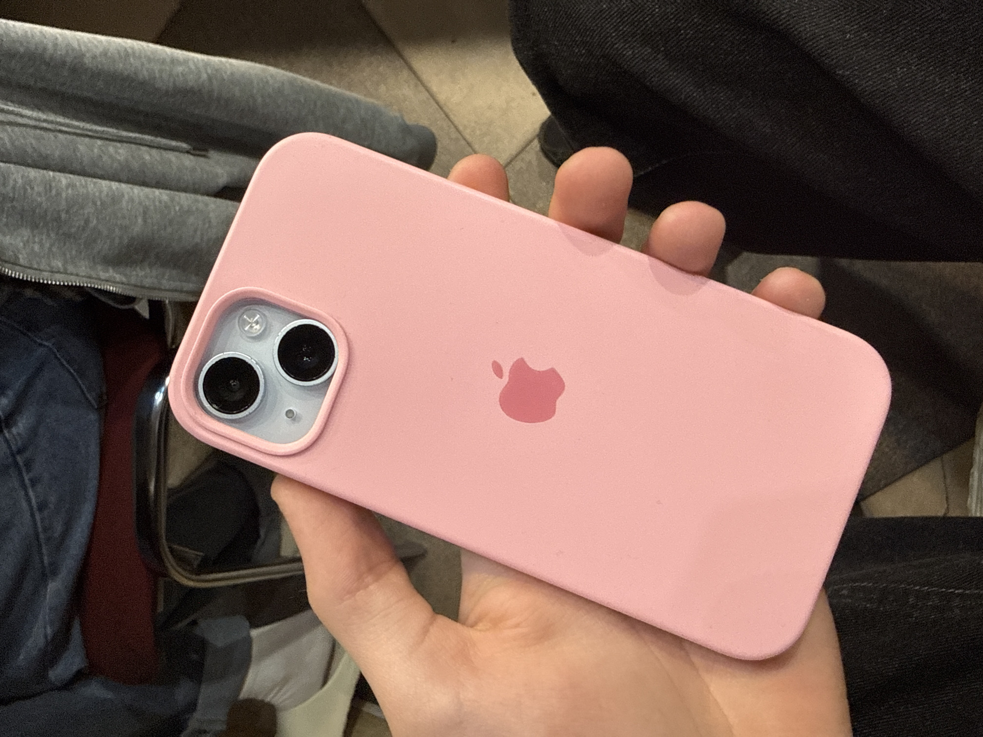 The image displays a close-up, slightly overhead view of a person's hand holding a pink iPhone, likely an iPhone 13 or 14, encased in a soft pink protective cover. The phone is oriented horizontally, showcasing its back. A dual-lens camera system is visible in the top-left corner of the phone's back, featuring two dark circular lenses within a slightly raised module, alongside a small flash and a possible sensor. The iconic pink Apple logo is centrally positioned on the phone's back.
The person's fingers gently grip the phone from below. In the blurred background, elements of an indoor setting are visible. To the left, a textured gray fabric, resembling a hooded sweatshirt with a zipper, is partially seen. To the right and below the phone, dark fabric, possibly jeans or trousers, is present. The floor appears to be tiled with light-colored square tiles. A metallic frame, possibly from furniture like a chair or table, is also discernible in the lower left portion of the background. The scene suggests a casual indoor environment, potentially in Birobidzhan, Russia. The time of day and weather cannot be determined from the available visual information. No specific activity or emotion is explicitly depicted beyond the act of holding the phone.