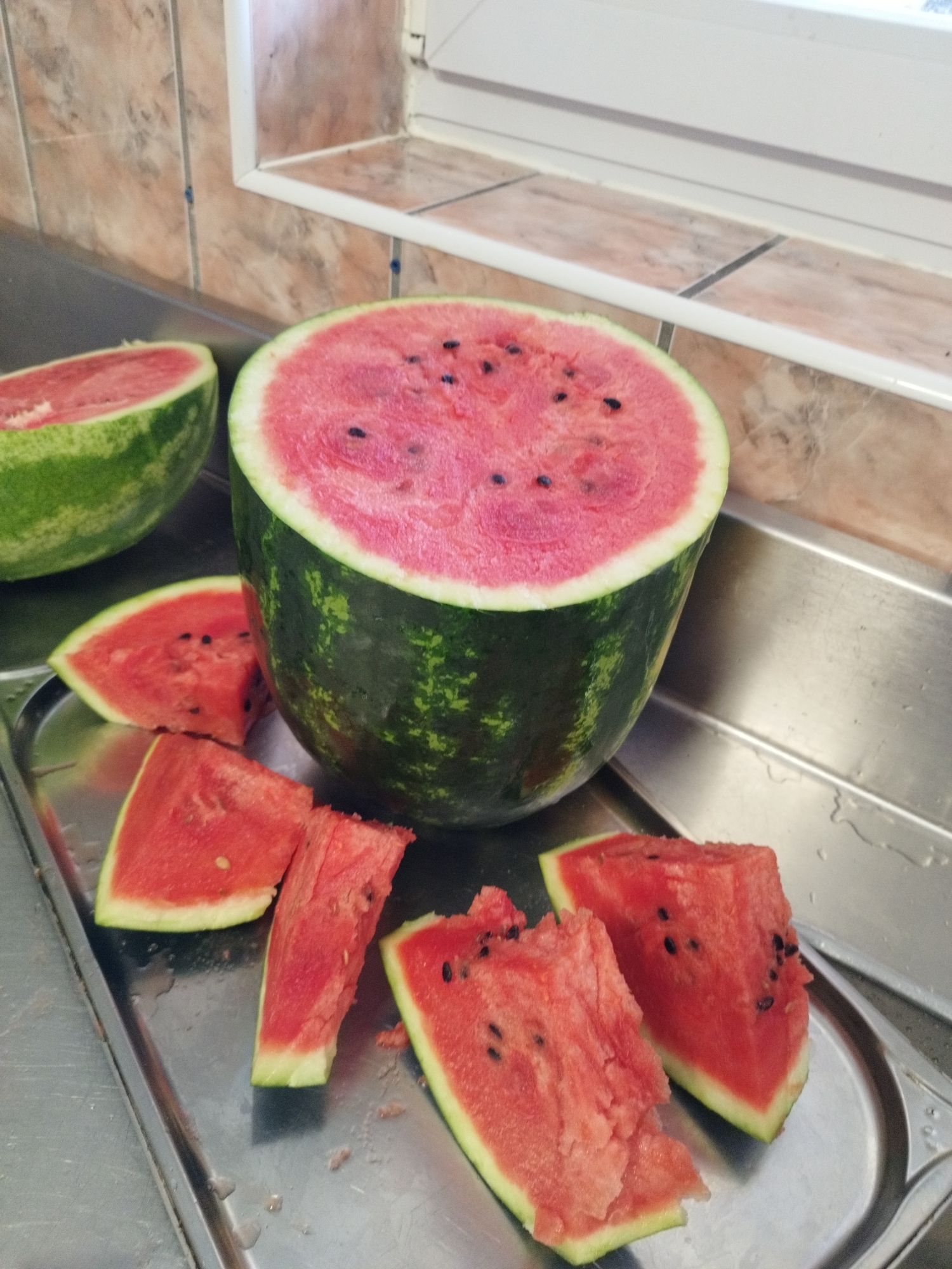 A halved watermelon rests on a stainless steel counter next to a partially sliced watermelon and several watermelon wedges. The halved watermelon is positioned upright, with its cut side facing the viewer, revealing its pink flesh and black seeds. The partially sliced watermelon is to the left, also cut side up. Several triangular watermelon wedges are arranged on a silver tray in the foreground. The background consists of a tiled wall and a window sill. The scene appears to be in a kitchen or food preparation area.