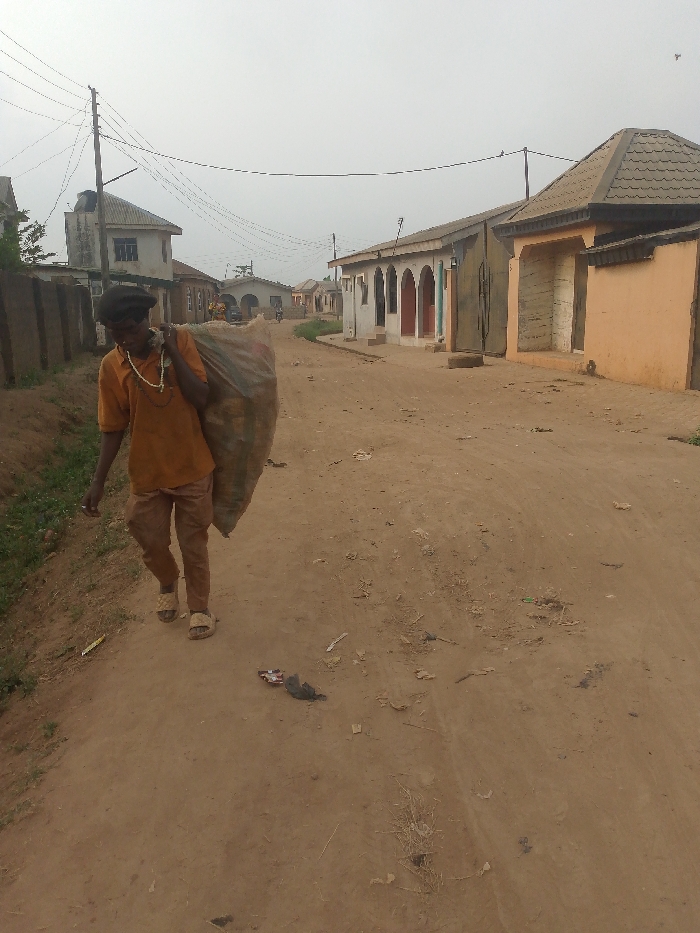 A man carries a large bag over his shoulder, walking along a dirt road in a village. He wears a yellow shirt, brown pants, and sandals. A string of beads is visible around his neck. The road is lined with simple houses of various colors. Overhead, power lines run along the street. Trash litters the road surface. The sky is overcast.