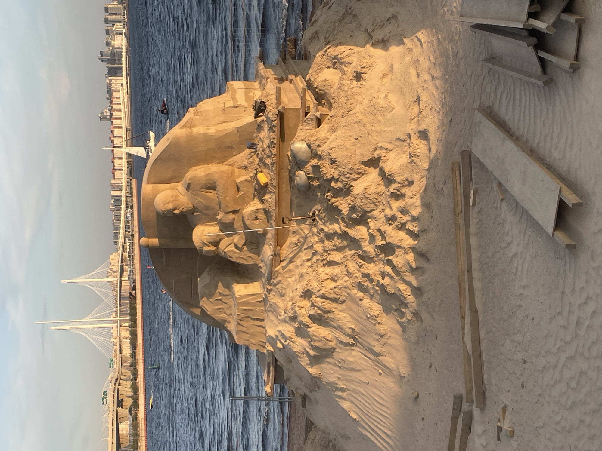 A sand sculpture depicts a man with a beard sitting and holding a young child on his lap. The sculpture is situated on a large mound of sand, with wooden planks scattered on the beach in the foreground. In the background, a body of water is visible, with a bridge, buildings, and a sailboat. The sky is clear with a few clouds.