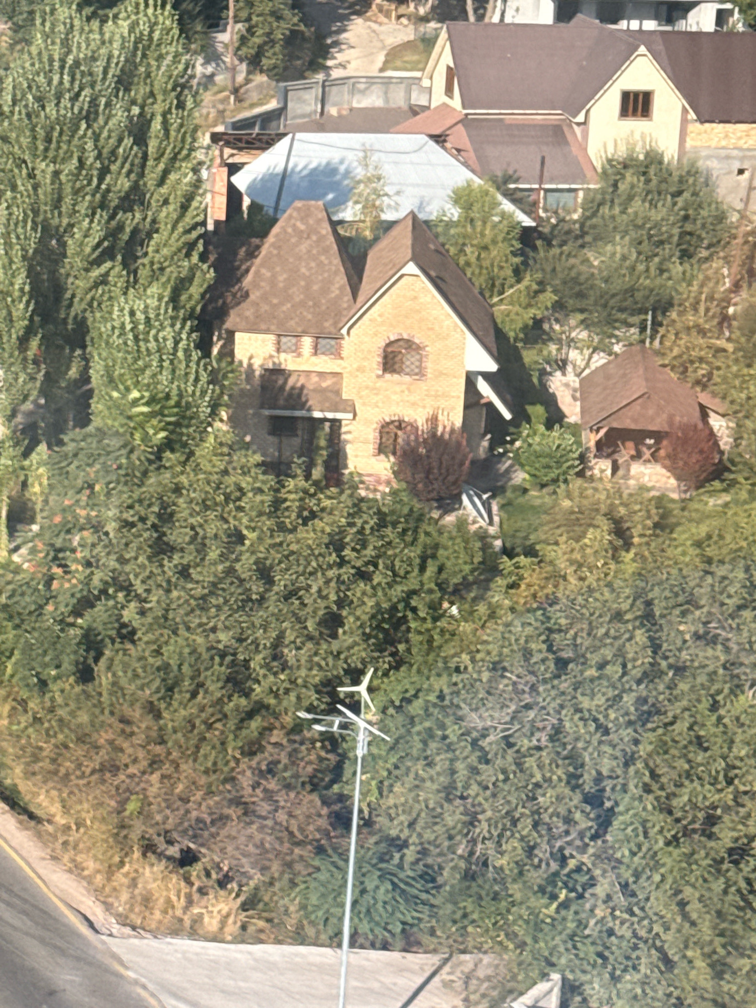 The image depicts a residential area, likely Amirsay, Uzbekistan, characterized by a cluster of houses nestled amidst lush greenery. In the foreground, a paved road curves along the left side of the frame, bordered by dry, yellowish grass. A tall, slender pole with a small wind turbine-like attachment and lights stands prominently in the lower right quadrant, casting a shadow.
Dominating the mid-ground is a two-story brick house with a steeply pitched, brown-tiled roof. The house features arched windows, some with visible panes, and a dark-colored entrance. Reddish-brown bushes are planted near its base. Adjacent to this house, and slightly behind it, is a small, brown, hexagonal wooden gazebo with a peaked roof, furnished with two chairs and a table. The surrounding landscape is densely covered with various trees and shrubs, creating a verdant and somewhat wild environment.
In the background, larger, more modern houses with different roof styles and lighter-colored facades are visible, suggesting a mix of architectural styles and stages of development within the community. Some of these structures are partially obscured by trees. The overall impression is of a tranquil, semi-rural residential setting.