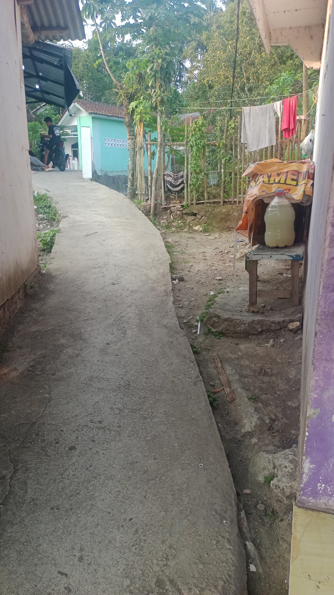 A narrow, paved pathway curves to the right, bordered by a textured wall on the left and uneven ground with sparse vegetation and debris on the right. In the background, a turquoise building with a patterned wall is visible, flanked by trees and vegetation. A clothesline with laundry hangs across the scene, and a large plastic bottle filled with a pale yellow liquid sits on a wooden stool. A young person is seated on a motorcycle in the upper left portion of the frame, and other people are indistinctly visible. The setting appears to be a residential area in Cogreg, Indonesia, with a mix of structures and natural elements.