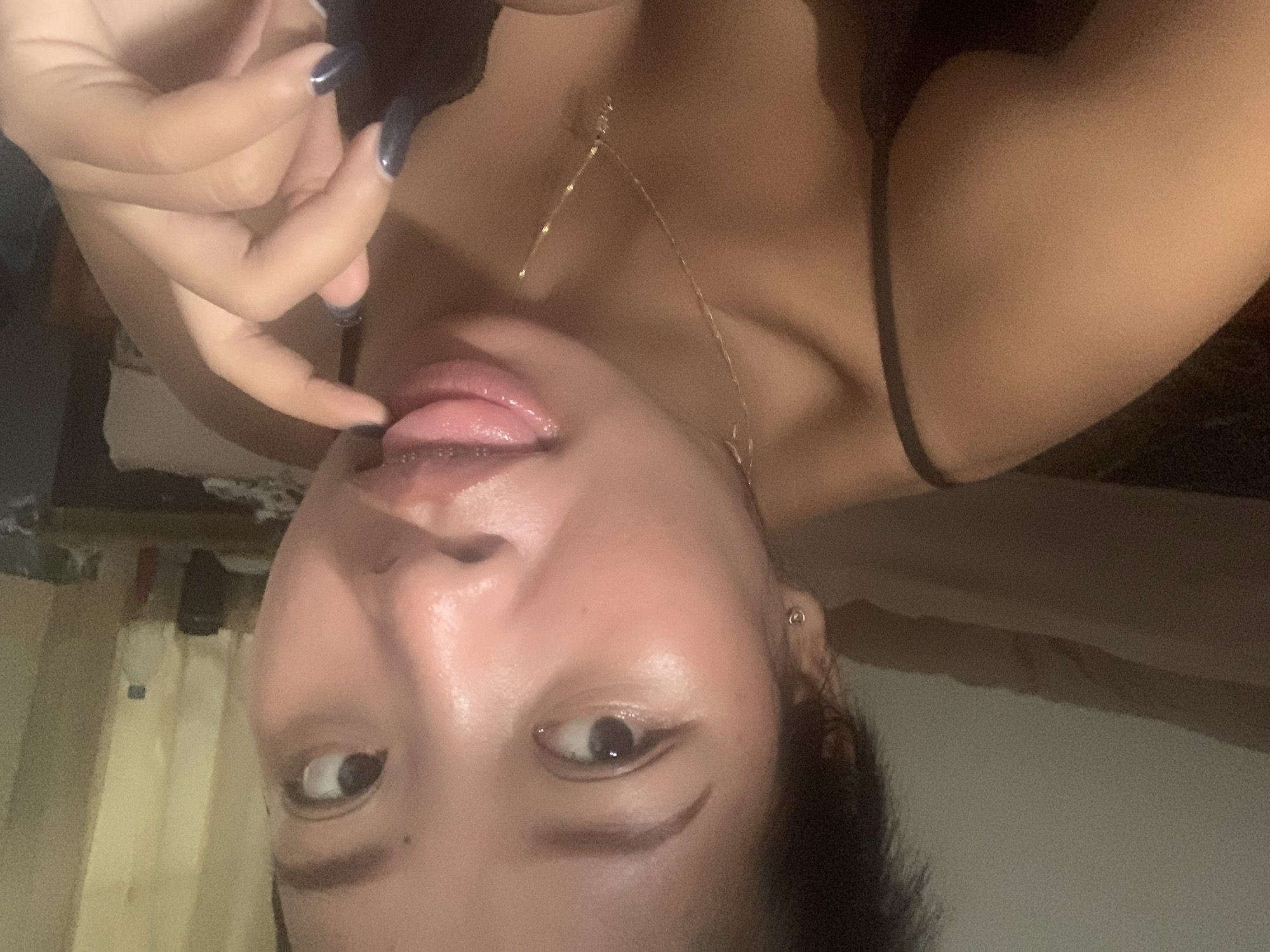 A person with dark hair is angled upside down with their finger touching their extended tongue. The person has dark, long nails and is wearing a thin gold necklace. The location is indistinct. There are objects that appear to be furniture and wall hangings in the background, but they are out of focus. The city of Denpasar, Indonesia, is not evident from the image.