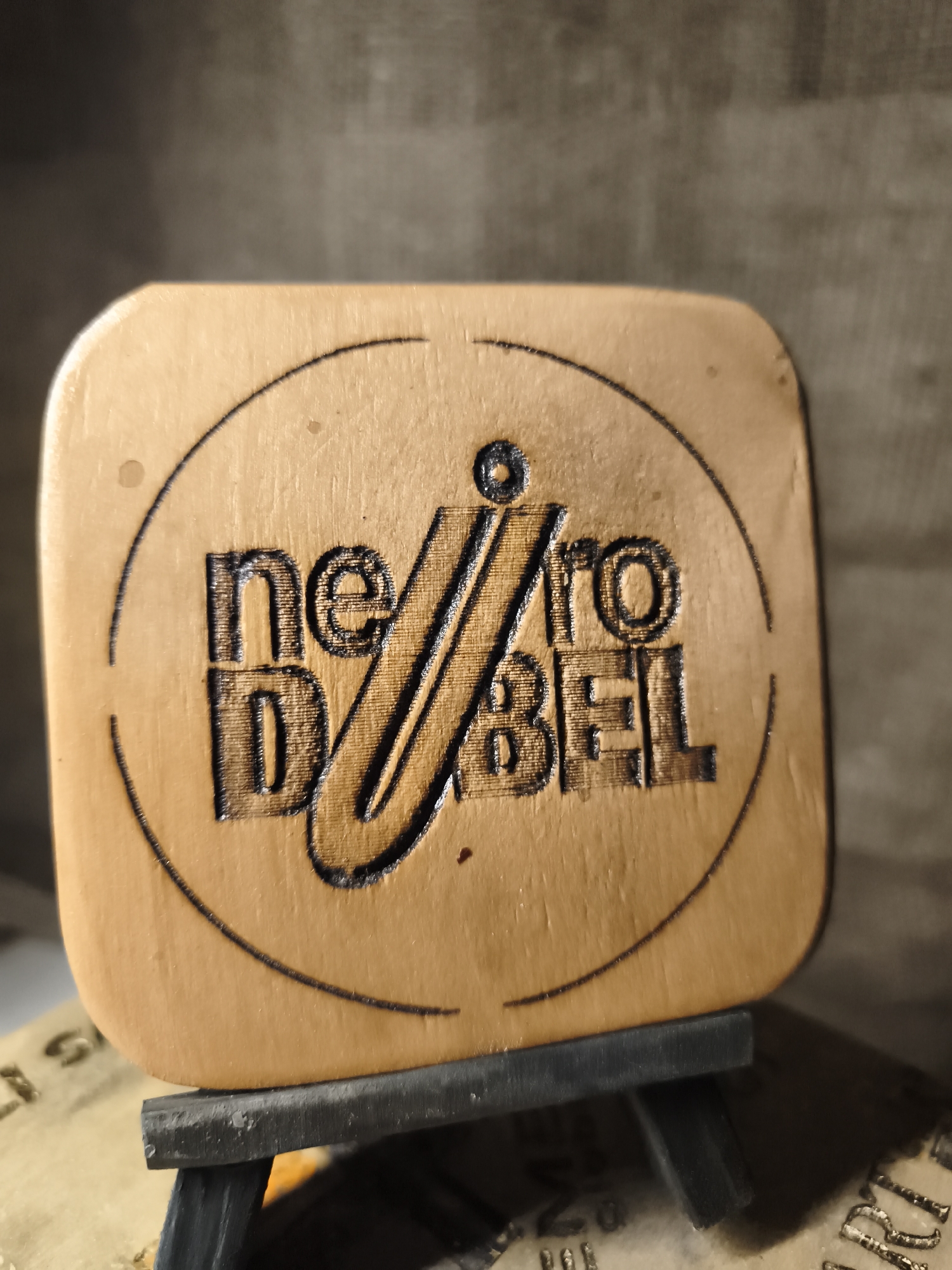 The image shows a close-up of a wooden object with a burnt-in logo. The logo is circular and contains text that appears to read "ne D BEL ro". The object is light brown and has a smooth, polished surface. It is resting on a small black easel, which is itself on top of a surface with some printed text. The background is a muted, textured gray. The scene is well-lit, with the light source casting a soft shadow on the right side of the object. The image is taken from a slightly elevated angle. There are no people visible in the image. The specific location in Гаранскі сельскі Савет, Belarus, is not discernible from the image.