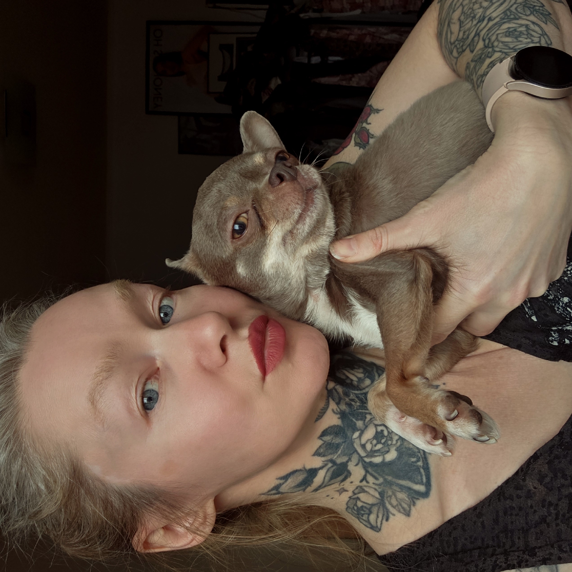A fair-skinned woman with blue eyes and blonde hair is holding a brown and white Chihuahua dog. The woman has tattoos on her neck and arms. She is wearing a black shirt and a rose-gold smart watch. The background is dark and blurry, but there appears to be a poster on the wall behind her. The location is not identifiable as Saint Petersburg, Russia.