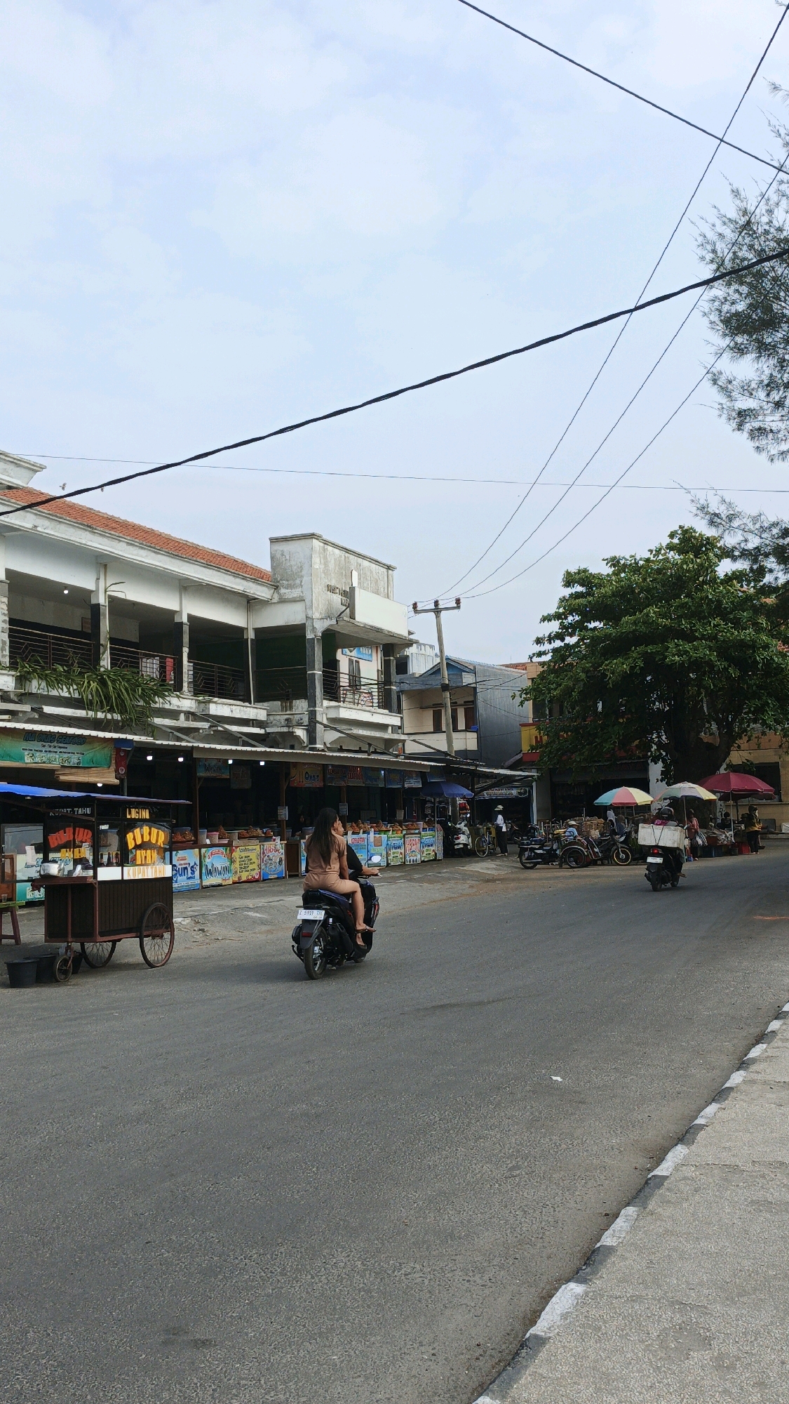 This is a medium shot, eye-level, exterior, vertical, daytime, full shot of a street in Desa Pagandaran, Indonesia. Two people, a man and a woman, are riding a black motorcycle down the street. The man is driving the motorcycle and the woman is sitting behind him, wearing a brown tank top and brown shorts. They are looking to the left. The street is paved and there are buildings on the left side of the street. The buildings have awnings and there are several food stalls set up in front of them. The food stalls are selling various items and there are signs in Indonesian. There are power lines running across the top of the image. On the right side of the street, there is a large tree, some umbrellas, and a few other motorcycles and scooters parked. The sky is cloudy.
