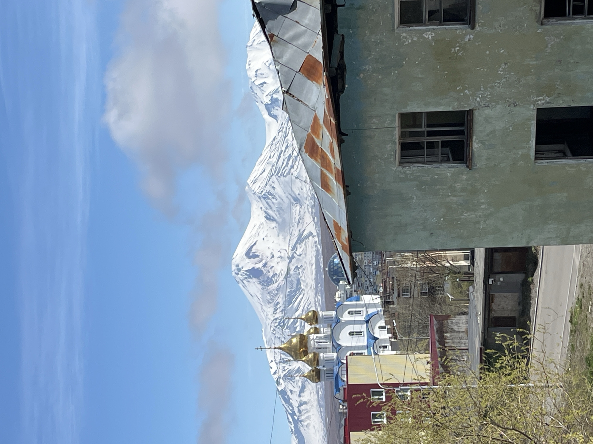 The image shows a view in Petropavlovsk-Kamchatsky, Russia. In the foreground, there's a dilapidated building with an aged, rusting roof. Its facade is a faded green, marked by empty window frames.
In the midground, a brightly colored Russian Orthodox Church with blue and white accents and gold domes stands out. Beyond the church, a massive, snow-covered mountain dominates the backdrop, its peak rising into a blue sky dotted with clouds.
The general scene evokes a contrast between urban decay and natural grandeur, highlighting the city's unique geographical setting.