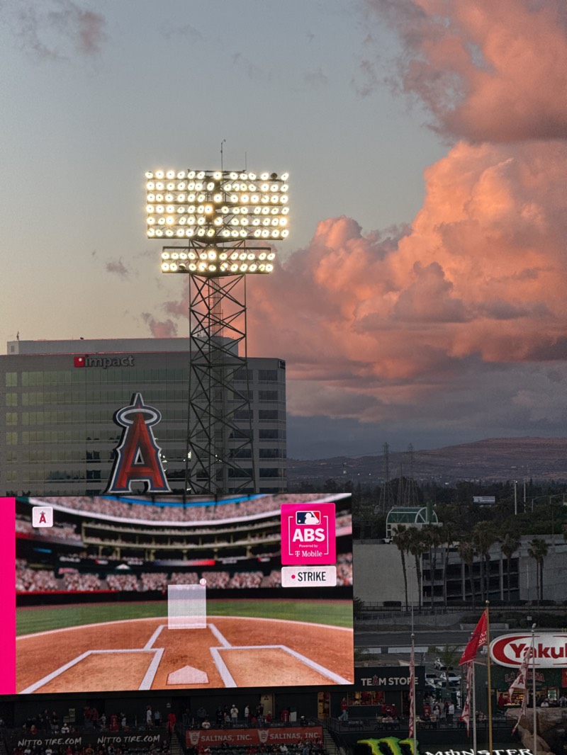 This media file captures an atmospheric evening view of a baseball stadium and its surroundings in Anaheim, United States, likely during an Angels game. The scene is dominated by a dramatic sky at dusk, featuring soft blue and pink hues blending into a vibrant orange as large, fluffy clouds catch the light of the setting sun.
In the foreground, a prominent stadium light tower stands tall, its numerous bright lamps illuminated, casting a warm glow. Behind it, a modern building with the "impact" logo is visible. A large, iconic red "A" logo, representing the Los Angeles Angels baseball team, is prominently displayed on another building, integrated into the stadium complex.
The lower half of the image features a large stadium scoreboard or jumbotron. It displays a graphic of a baseball field from the catcher's perspective, showing a pitch in motion. Text on the scoreboard reads "ABS powered by T-Mobile" and "• STRIKE", indicating an automated ball-strike system or a game replay feature. Below the scoreboard, actual stadium seating areas are visible, with small figures of spectators in the stands. Various advertisements are also visible on the stadium structures, including "NITTO TIRE.COM" and "SALINAS, INC.".
In the mid-ground and background, the landscape includes other buildings, clusters of palm trees, and distant hills or mountains under the colorful sky. Towards the bottom right, flags or banners with the "Yakult" logo and a partial "Monster" (Energy Drink) logo are seen, along with a sign that appears to say "TEAM STOPE" (likely "TEAM STORE"). The overall impression is one of an active evening at a major league baseball venue, enhanced by the picturesque sunset.