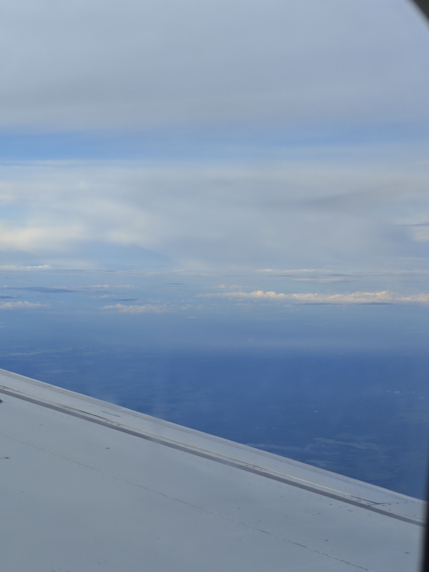 An aerial view from an airplane shows a bright, overcast sky with scattered clouds over a deep blue expanse, possibly the ocean or a large body of water. The lower portion of the image is dominated by the wing of the aircraft, which is white and shows faint lines and a few small dark spots, possibly debris. The horizon line is indistinct, blurred by the atmospheric conditions. The overall scene conveys a sense of height and distance, with a muted color palette of blues and whites.