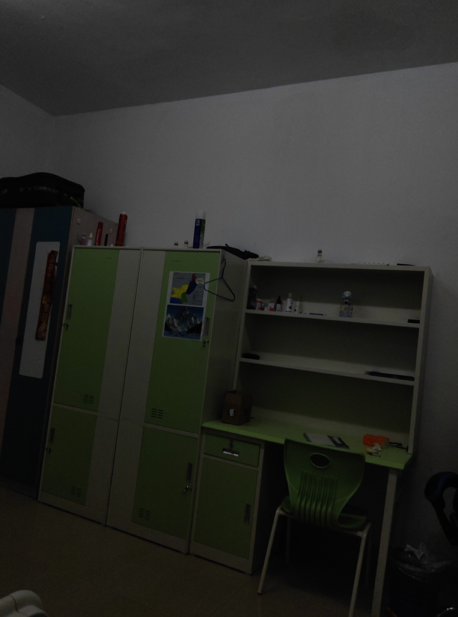 The image depicts an indoor scene, likely a dormitory or study room, in Chongqing High-tech Zone, China. The room contains a large green and white wardrobe on the left, a white bookshelf with three shelves on the right, and a green desk with a chair in front of it. Several items are visible on the shelves and desk, including bottles, what appears to be a trophy, and papers. The lighting is dim, suggesting it might be evening or the room's lights are not fully on. The overall impression is one of a functional, yet somewhat cluttered, personal space.