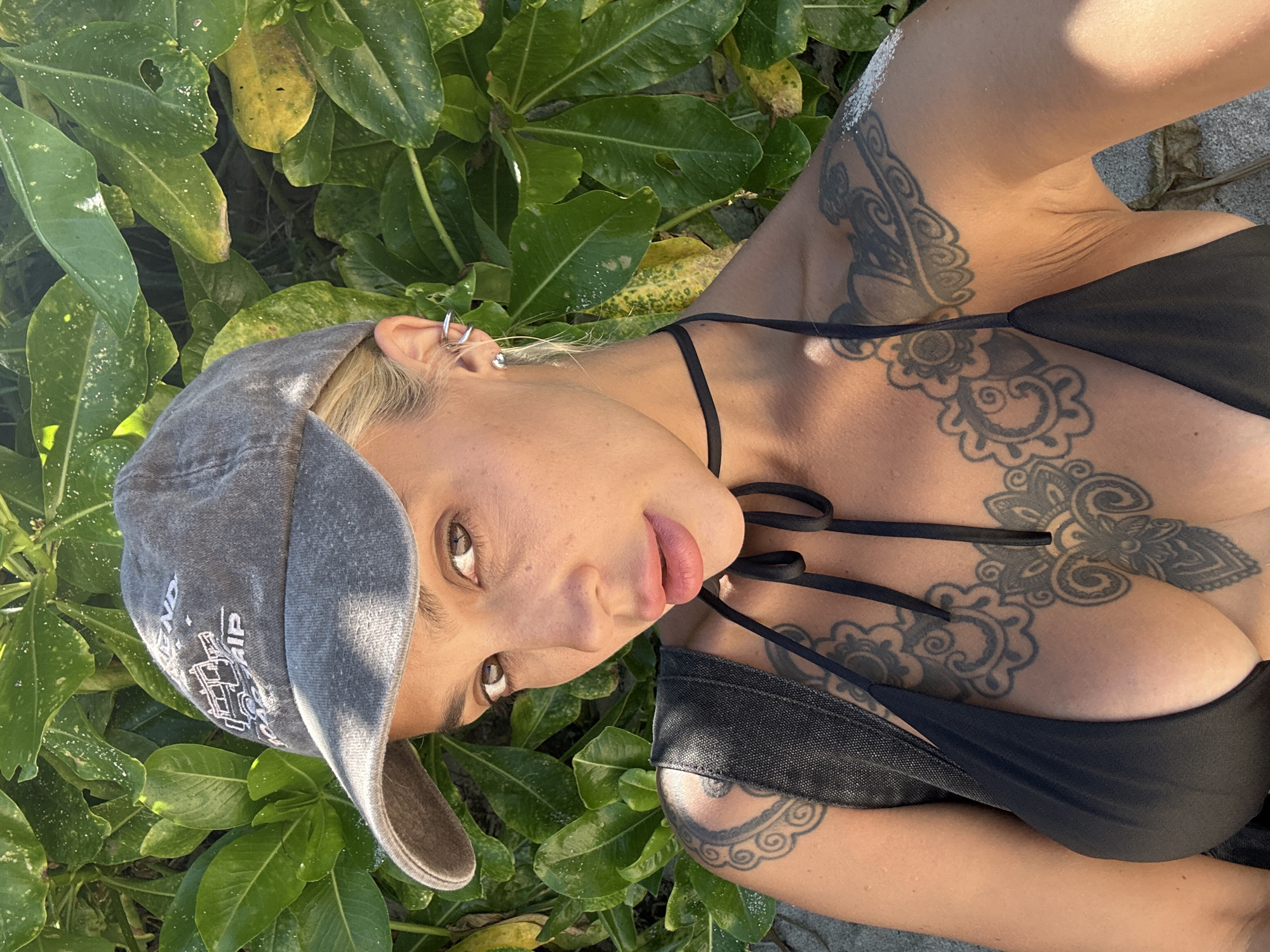 A woman with blonde hair, wearing a grey baseball cap and a black bikini top, is lying down amongst green foliage. She has tattoos on her chest, arm, and shoulder. Her face is turned towards the camera, and she appears to be looking directly at the viewer. Her lips are slightly parted, and she has a casual expression. The lighting suggests it is daytime, likely with bright sunlight. The green leaves and sandy ground indicate an outdoor, possibly tropical, setting. The text on her cap is not clearly legible. The image is taken from a high-angle perspective, looking down at the woman.