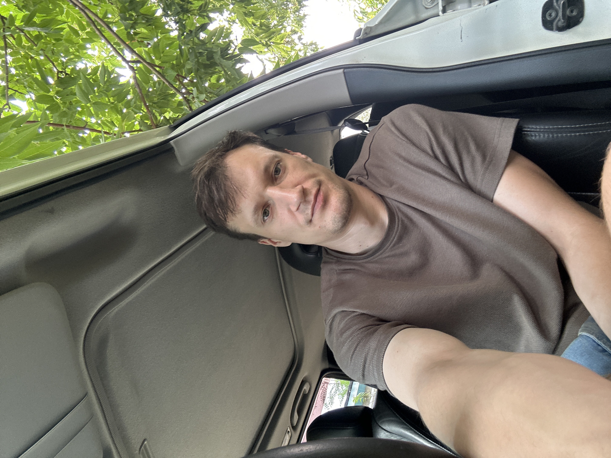 A man is sitting in the driver's seat of a car, taking a selfie. He has short brown hair and is wearing a brown t-shirt and blue jeans. The car's interior is visible, including the sunroof and a portion of the dashboard. Outside the car, lush green foliage and trees are seen. The scene appears to be in a natural, possibly rural or suburban, setting, with no specific urban landmarks of Makiv, Ukraine, being identifiable.