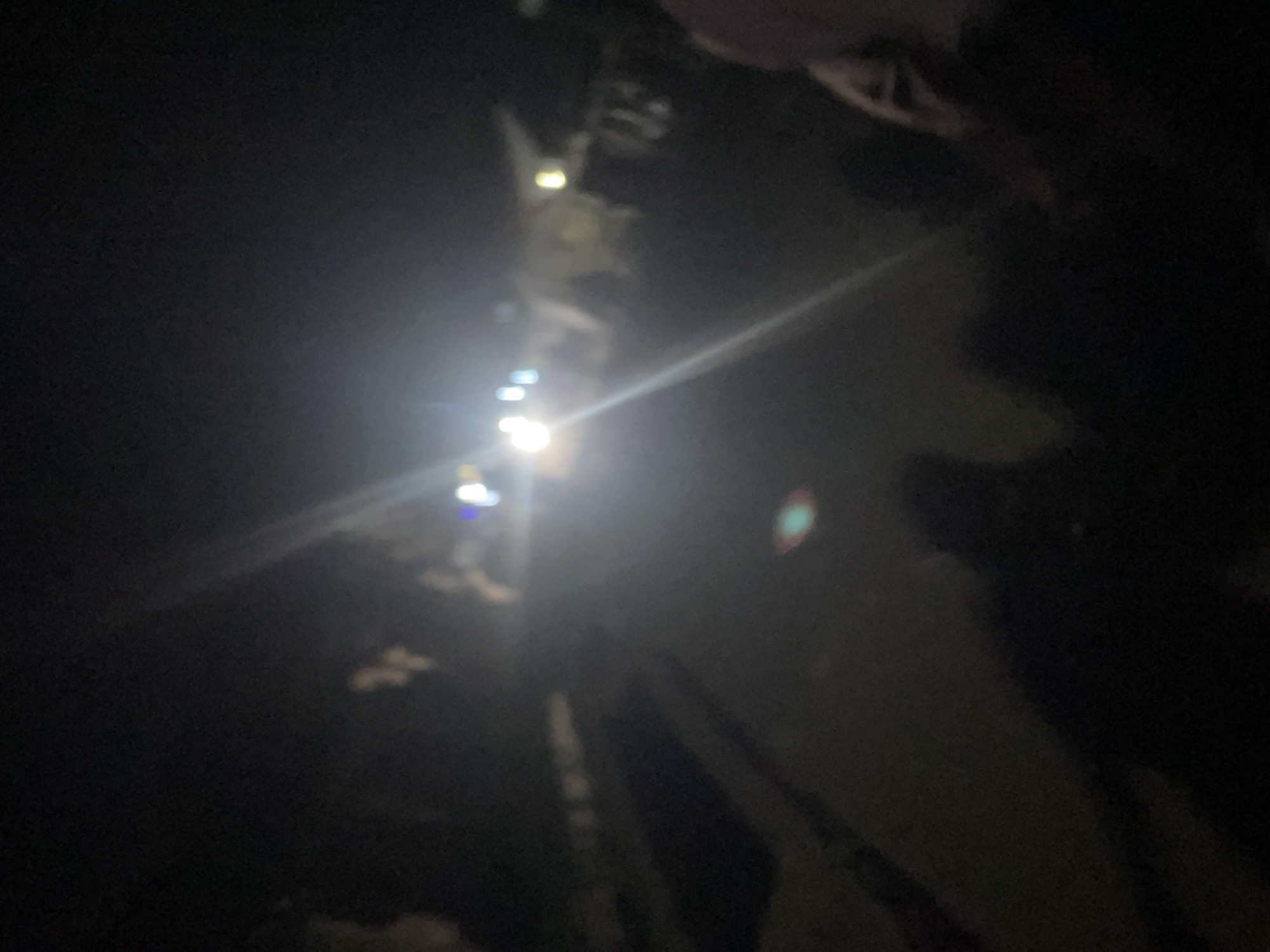 The image depicts a dimly lit scene at night, likely in Lokoja, Nigeria, as suggested by the prompt's context. The primary focus is a road or pathway illuminated by bright lights, possibly from vehicles or streetlights, creating lens flares and glare that obscure finer details. Several blurry figures are present, suggesting people are in the vicinity. Some appear to be standing or walking along the path, while others are closer to the foreground, also indistinct. The overall impression is one of movement and activity in a low-light urban environment.