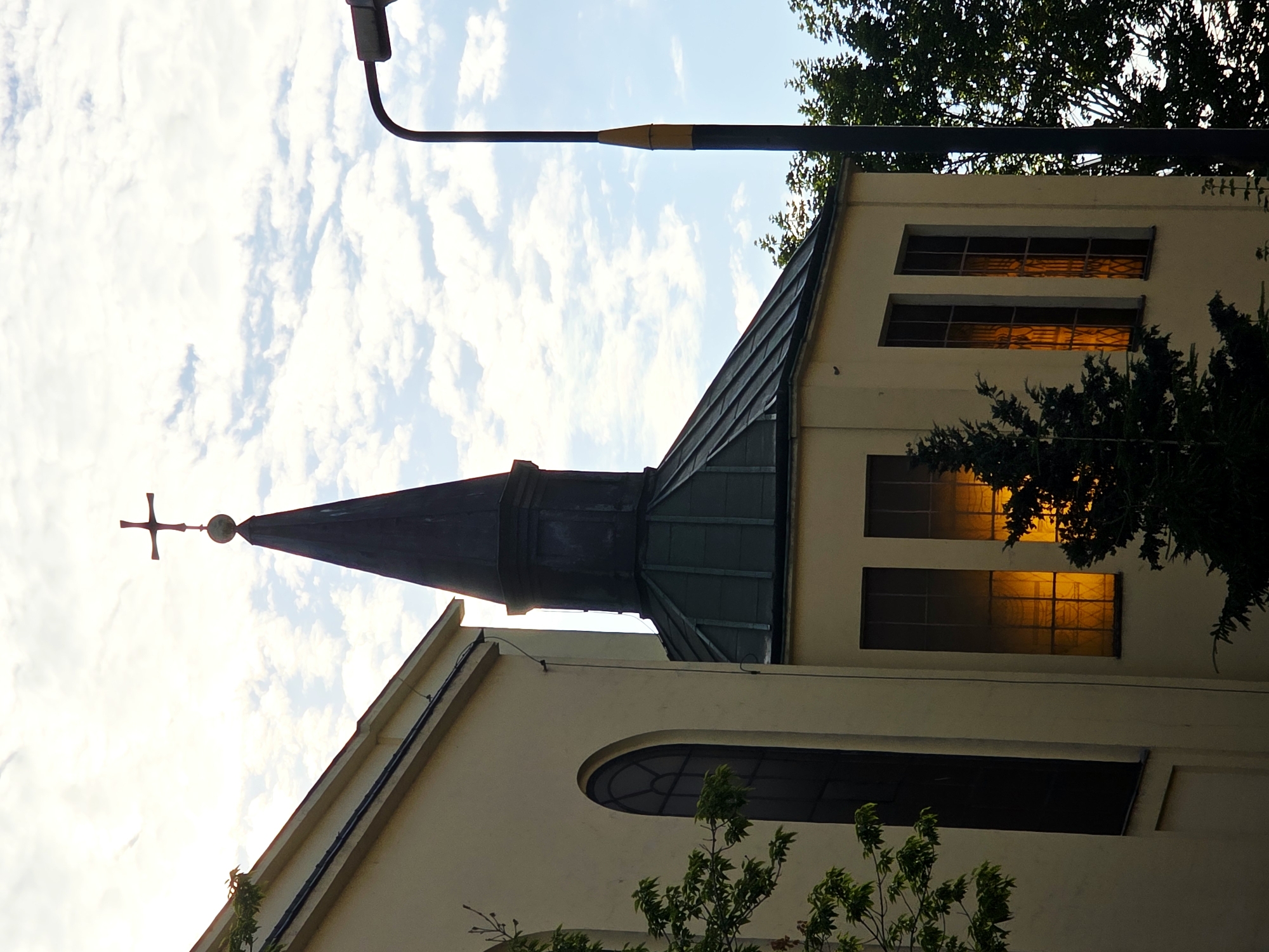 The image shows a church tower topped with a cross, set against a cloudy sky. The church building is cream-colored, with a large arched window on the left side and several tall, narrow windows on the right side, which are illuminated with a warm, orange light from within. A dark green, conical roof section extends from the tower. A tall, black lamppost with yellow accents stands to the right of the church, and trees are visible on the right and left sides of the frame. The lighting suggests it might be late afternoon or early evening. The scene is devoid of any people or visible activity, presenting a still and architectural composition. This image likely depicts St. Andrew Bobola's Church located at Rakowiecka 61 in Warsaw, Poland, which features a prominent tower and similar architectural style.