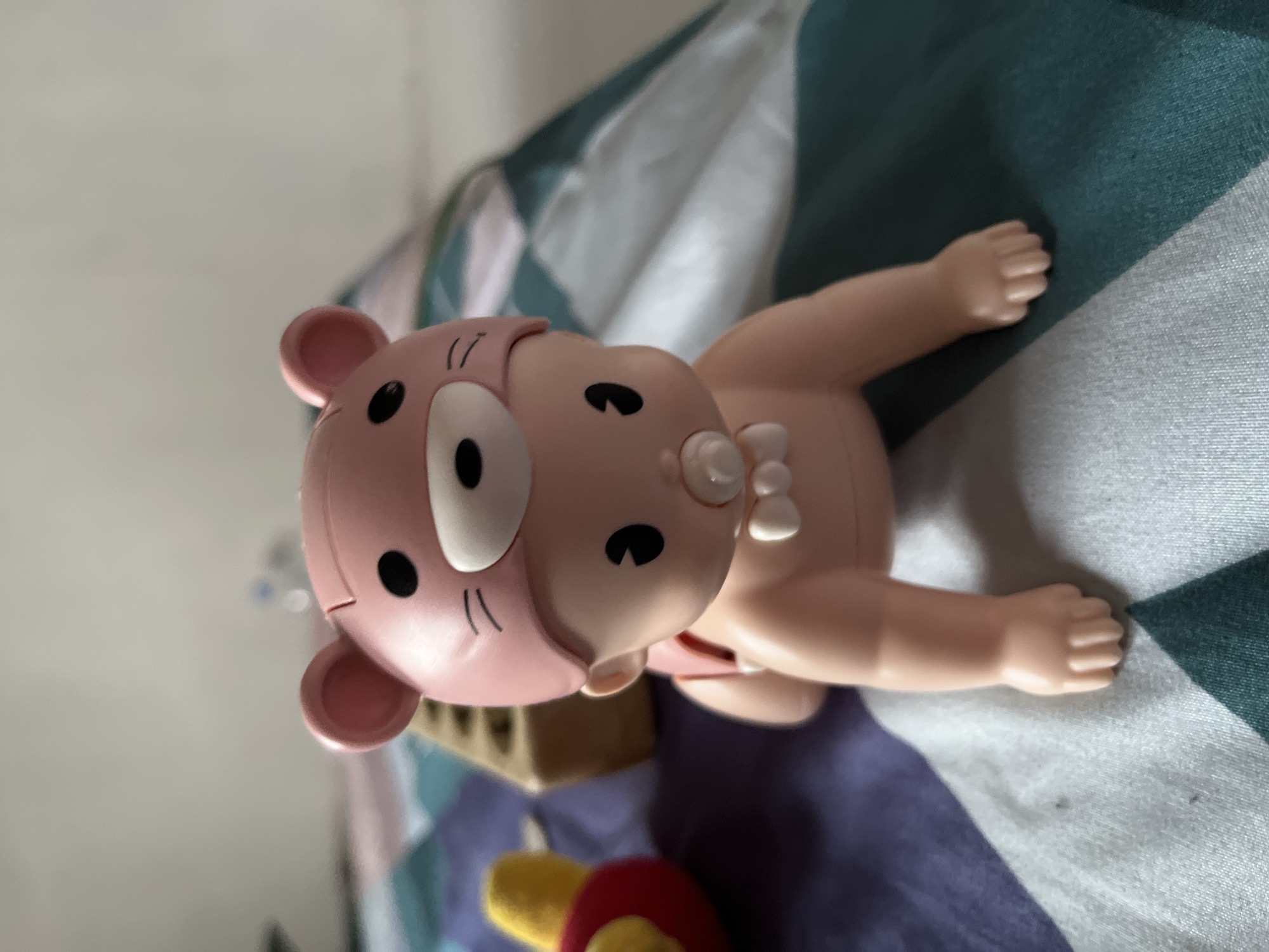 A close-up, eye-level, medium shot captures a pink baby doll wearing a bear-shaped hat. The doll is in a crawling position on a patterned blanket, with its right arm and leg extended forward. The blanket features stripes of teal, purple, and white, with subtle variations in pattern. The background is blurred, showing a light-colored wall and an out-of-focus object to the left. The lighting is soft, casting a slight shadow on the right side of the doll. There is no discernible city or specific location in Lhokseumawe, Indonesia, depicted in this image.