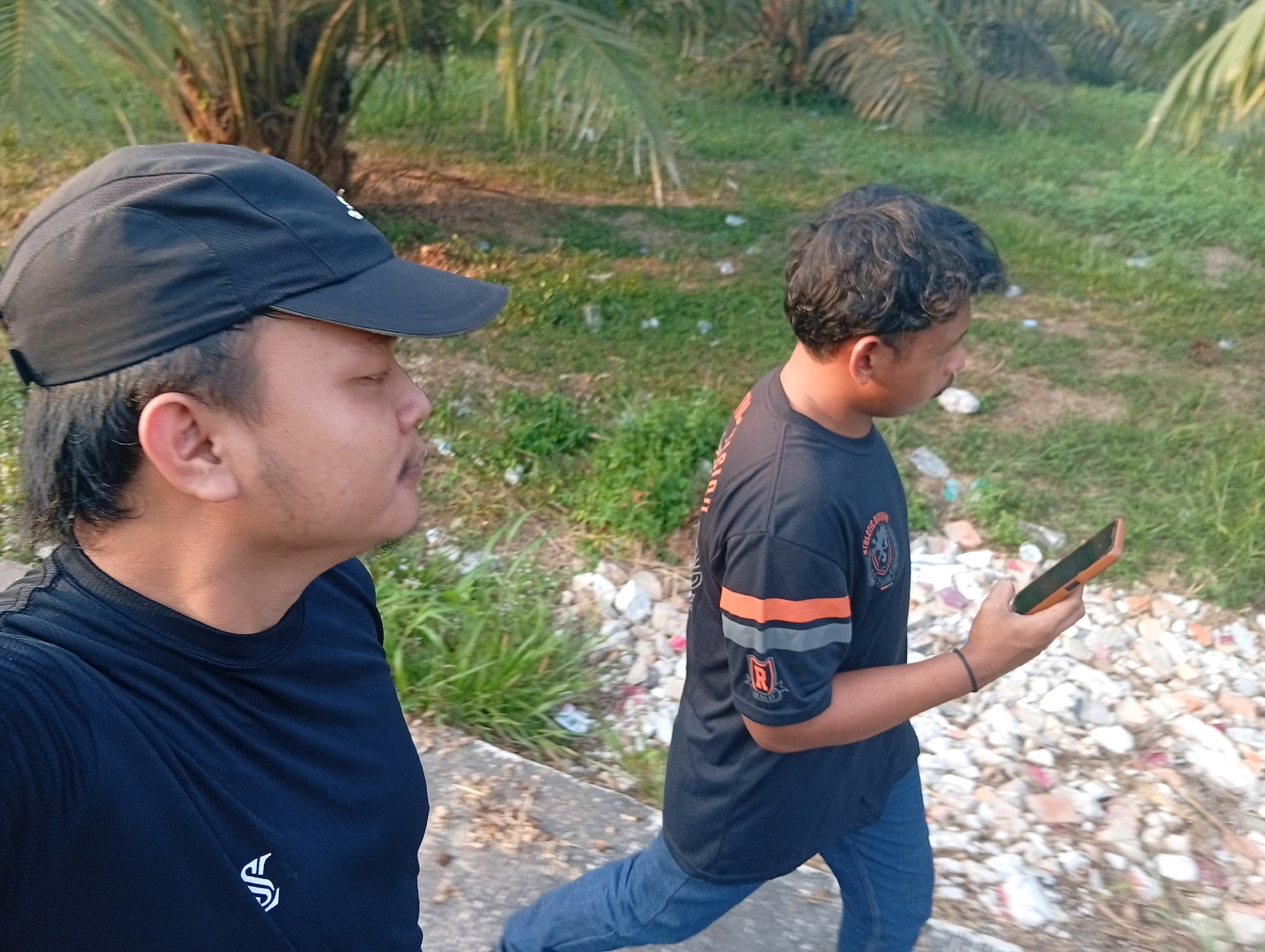 Two individuals are captured in a sideways, medium shot, with the foreground subject on the left, facing right, and the background subject on the right, also facing right. The foreground individual wears a black baseball cap and a black t-shirt with a white emblem on the left chest. The background individual is dressed in a black t-shirt with orange and grey stripes on the sleeves and a circular emblem on the left chest. This individual is holding a mobile phone in their right hand and appears to be walking on a pathway. The environment suggests an outdoor setting with lush green grass and palm trees visible in the background, hinting at a tropical or subtropical climate. The pathway is made of concrete, with scattered debris, including white and pinkish pieces, alongside the path. The lighting indicates it is daytime, possibly late afternoon, with a warm glow on the scene.