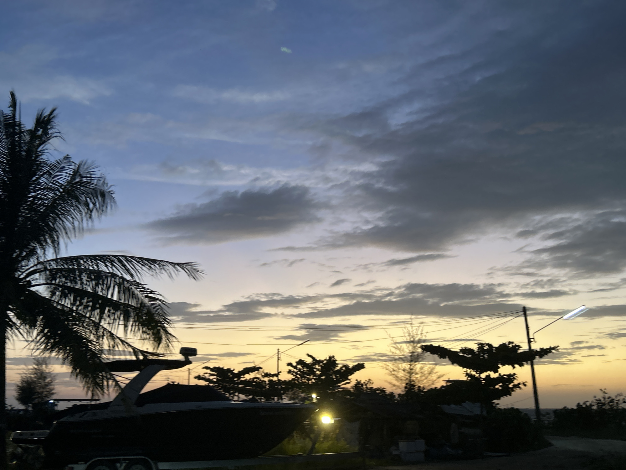 The image depicts a silhouetted scene at dusk in the Ko Pha-ngan District of Thailand. In the foreground, a dark-colored boat on a trailer is partially visible on the left. To the left of the boat, the fronds of a palm tree are silhouetted against the sky. Behind the boat and trees, a streetlamp illuminates a brightly lit, rectangular fixture. The sky transitions from a pale yellow near the horizon to a deep blue at the top, with scattered clouds. Power lines are visible stretching across the scene, supported by utility poles. The background shows a silhouette of trees and a few structures, suggesting a coastal or semi-rural area. The overall impression is one of twilight settling over a tropical landscape.