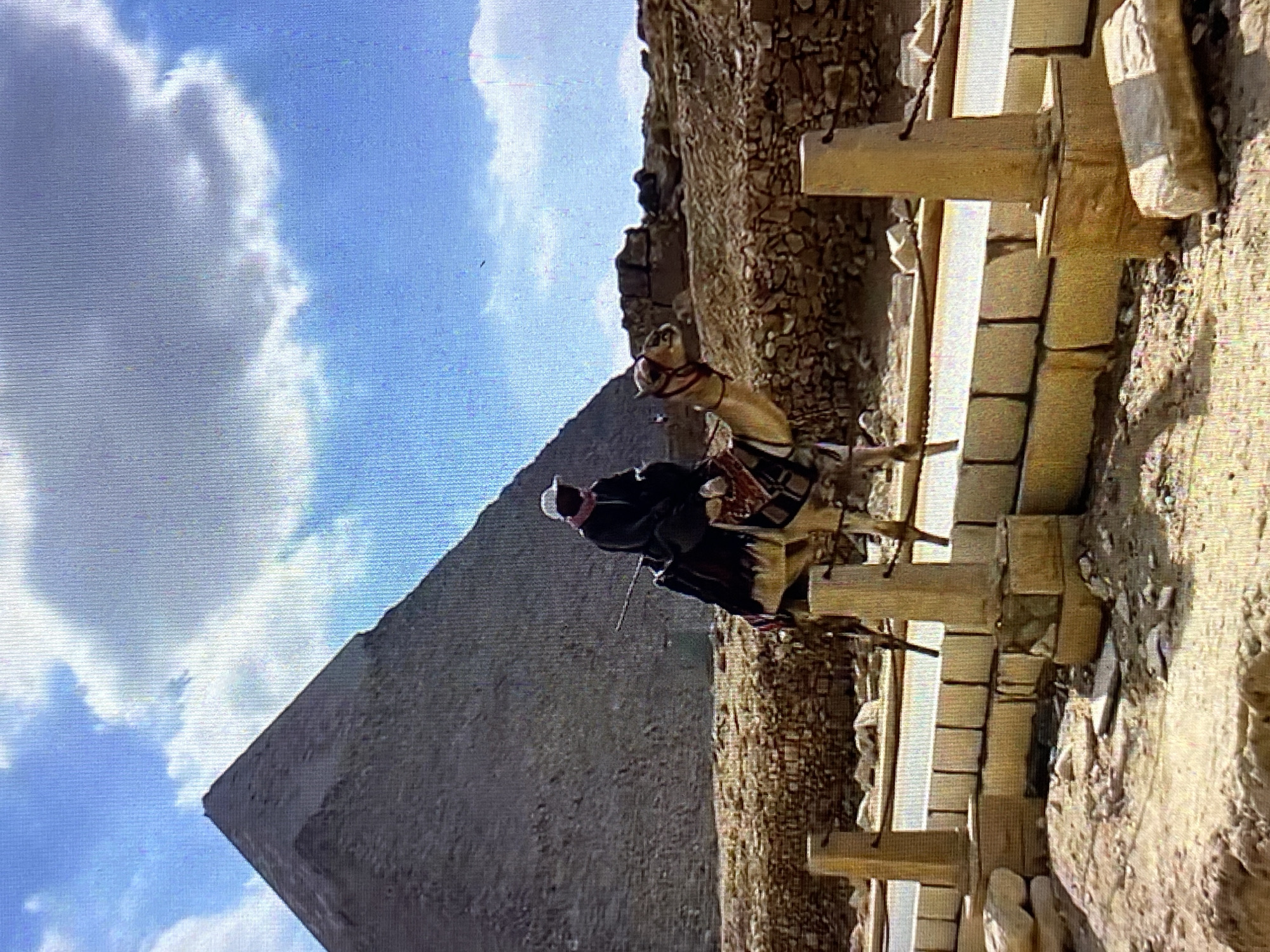 The image features a pyramid against a blue sky with clouds. A man wearing a head covering and dark clothing is riding a camel. The scene is partially obstructed by a series of tan rectangular blocks and weathered stone structures. The location is near a pyramid, suggesting it might be in Giza, Egypt, rather than Tinaquillo, Venezuela.
