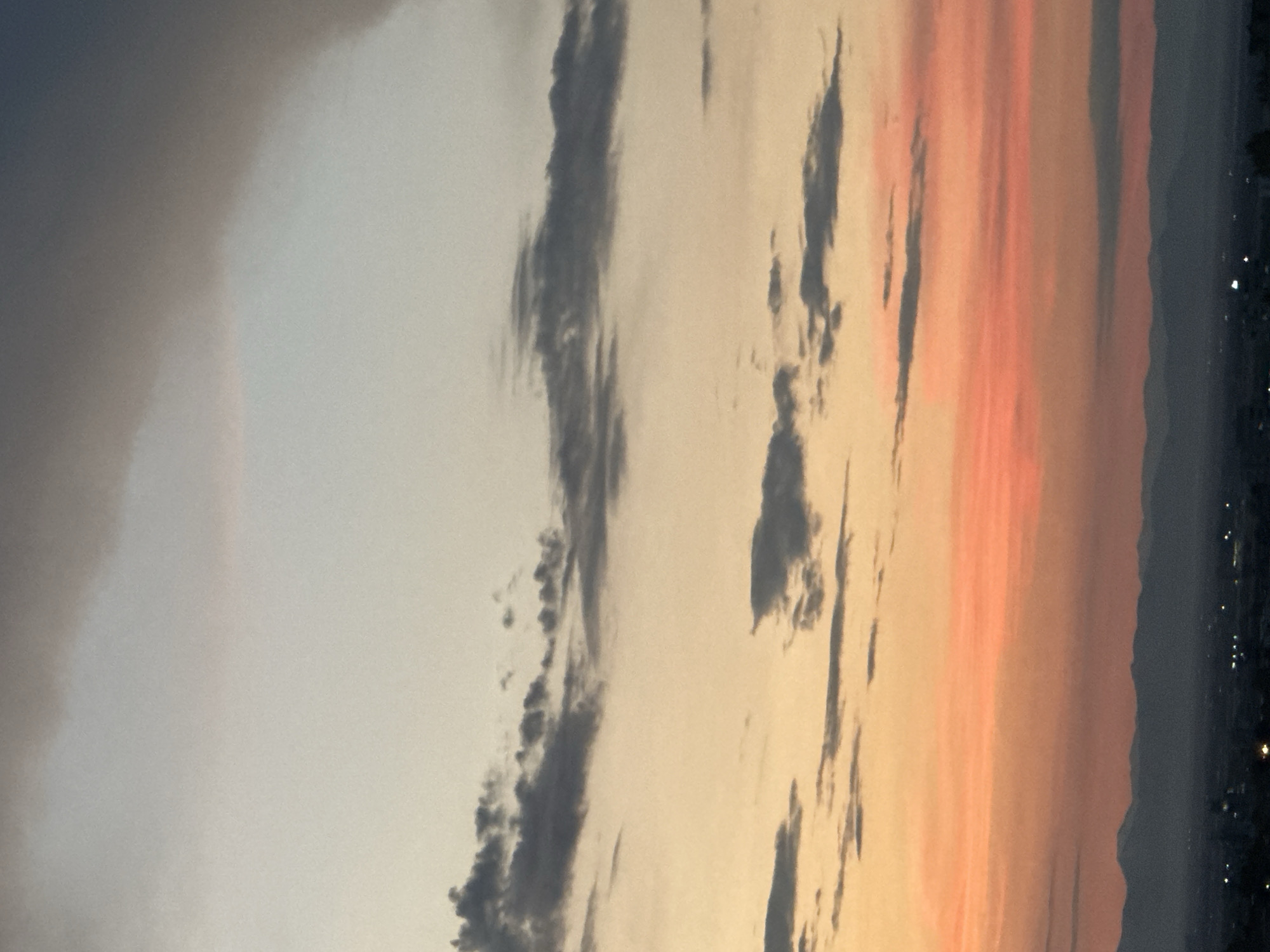 The image captures a panoramic view of Asenovgrad, Bulgaria, during sunset. The sky is a canvas of soft hues, transitioning from a pale blue at the top to a vibrant orange and pink near the horizon. Wispy clouds are scattered across the sky, with darker clouds forming a band across the upper portion of the image. In the distance, a silhouette of mountains can be seen against the colorful sky, with the faint outlines of buildings and lights of the city of Asenovgrad visible at the bottom. The scene is devoid of any people or specific actions, presenting a serene and atmospheric depiction of a sunset over a Bulgarian city.