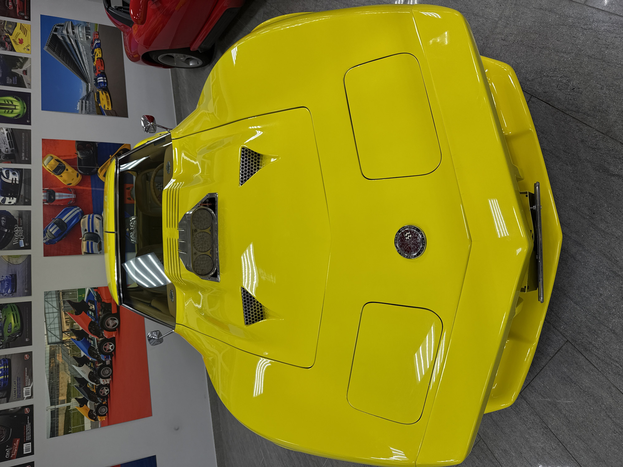 The image presents a bright yellow vintage sports car, likely a Chevrolet Corvette C3 Stingray, viewed from an overhead perspective within an indoor setting in Sirius, Russia. The car's glossy finish reflects bright overhead lights, highlighting its sculpted body. Notable features on the yellow car include a prominent hood scoop with visible air intakes, two triangular mesh vents on the hood, and covers for pop-up headlights. A circular emblem, characteristic of a Corvette badge, is centered on the front nose. Chrome side mirrors are also visible.
The car is parked on a dark gray floor, possibly tiled or concrete. In the upper right corner, a partial view of a red car is visible, suggesting other vehicles are present in the same location. The white walls surrounding the car are decorated with numerous framed posters and images, primarily featuring other sports cars. Two large posters are prominent: one shows a vertical arrangement of colorful cars against a modern architectural background, and another displays several sports cars lined up on a track from an elevated viewpoint. Smaller posters also depict various cars, with fragmented text such as "ROAD TRIP," "CHALLENGE," "Vegas," "Loader Car," and "GEN7" being partially discernible. One small poster features a Canadian flag motif. The scene is a static display, characteristic of a showroom, museum, or private collection, and there is no activity taking place other than observation of the vehicles. The lighting is artificial and uniform.