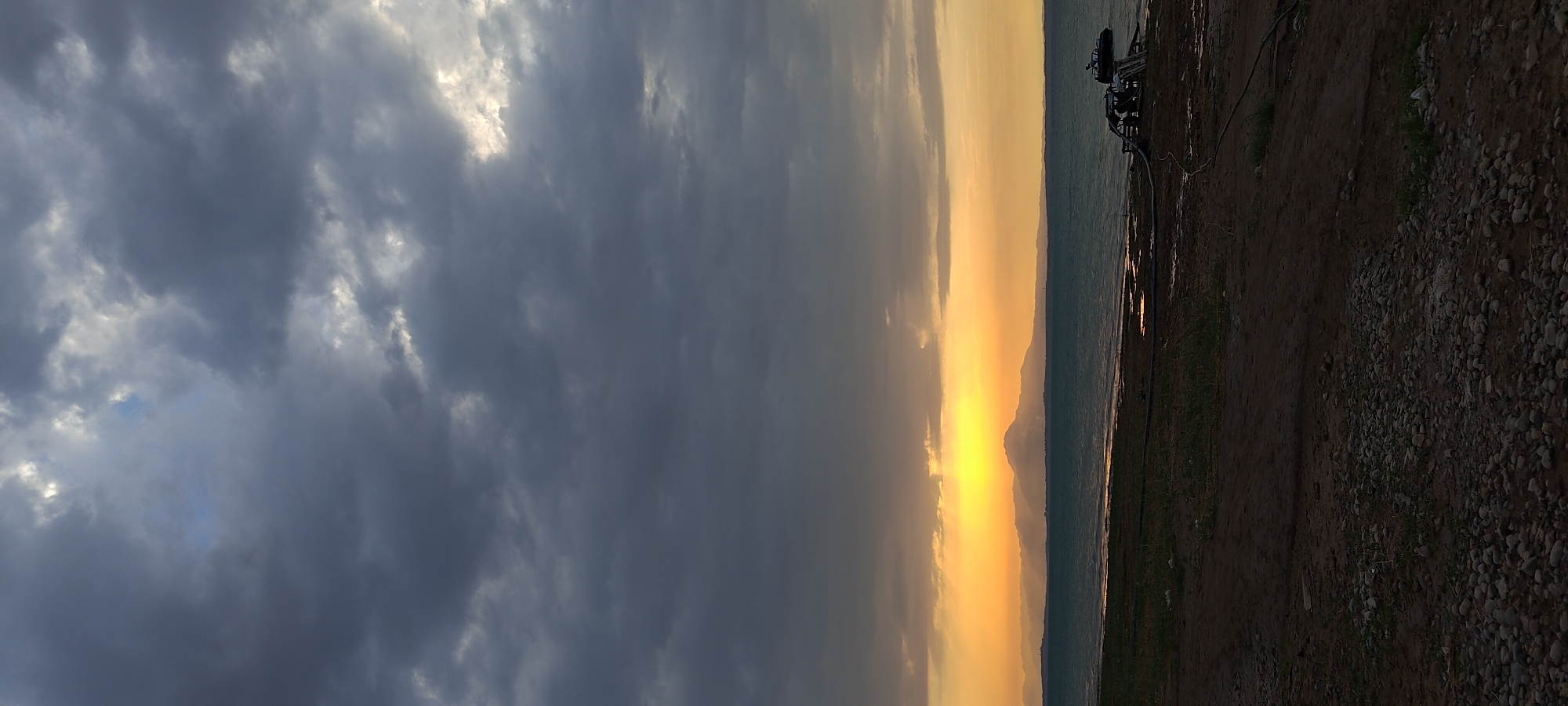 The image depicts a sunset over a body of water, with a mountain range in the distance. The sky is filled with dramatic, dark clouds, with a bright patch of sunlight breaking through on the horizon. The water is choppy, and there is a small, dark object that appears to be a boat or raft on the right side of the frame. The foreground consists of a rough, pebbly ground with some sparse vegetation. The scene is likely set in Chigdalik, Tajikistan, given the mention of the location. There are no people visible in the image.