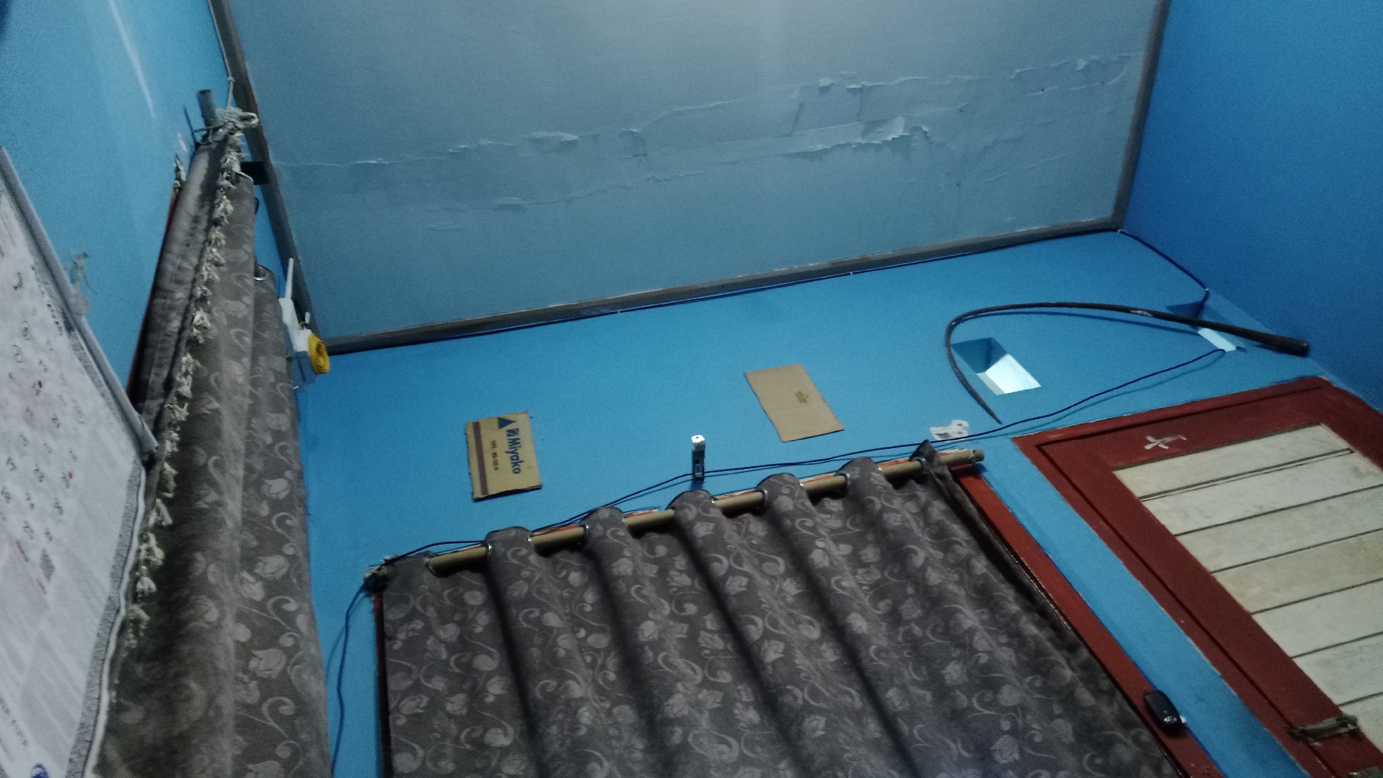 The image shows a corner of a room with blue walls and a light blue ceiling that appears to be peeling in places. On the left, a portion of a calendar or large poster with text and some drawings is visible. A gray curtain with a floral pattern hangs from a rod across the middle of the image, partially obscuring the wall.
On the blue wall, there are several objects. To the left of the curtain, a white and yellow object, possibly a Wi-Fi router or power adapter, is mounted on the wall. To the right of the curtain, there are two small cardboard pieces attached to the wall. The one on the left has the text "Miyako" in black and yellow lettering. Above the curtain rod, there is a black cord that snakes across the wall.
In the upper right portion of the image, a dark, whip-like object is draped over the wall, with its tip resting near the ceiling. Below this, a framed window or cabinet with a dark red frame is visible. The window itself has horizontal slats, some of which are white. A small black electrical switch or outlet is attached to the wall near the window frame.
The scene appears to be an indoor residential space, likely a bedroom or living area. The lighting suggests it is either nighttime or the room is dimly lit. Given the presence of the calendar and the general decor, this could be a typical room in a home in Denpasar, Indonesia, a city known for its tropical climate and residential areas.