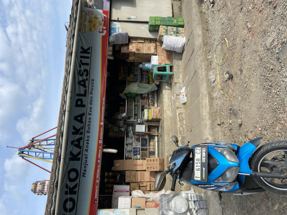 The image shows a storefront in Sukatani, Indonesia, located at coordinates -6.611 latitude and 107.412 longitude. The store is called "Toko Kaka Plastik" and sells various baking supplies and plastic goods. There is a blue scooter parked in front of the store. In the background, a large, colorful Ferris wheel is visible. The store is open with shelves full of various products. A person is standing near the entrance of the store. A person is also standing near the store entrance, possibly a customer. The store has a red and white awning with the store name on it. A few items, including a plastic chair, are on the sidewalk in front of the store. The ground in front of the store is covered with small gravel. The sky above the store is mostly clear with a few clouds. The scene appears to be a small local business district with a street fair or carnival nearby.