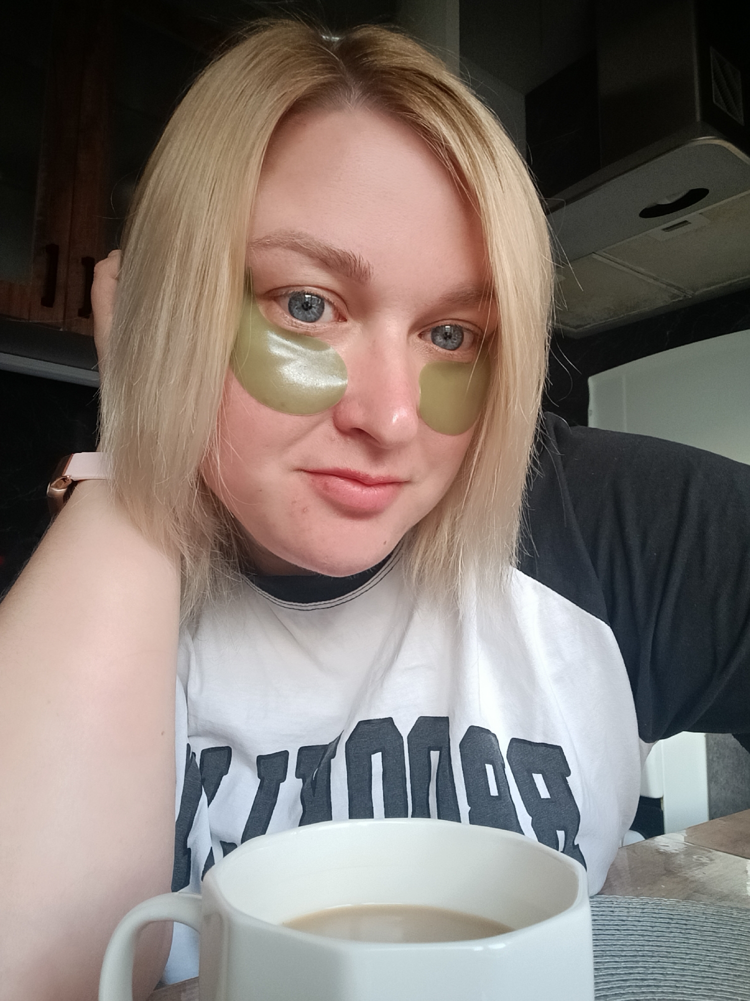 A blonde woman with blue eyes, wearing muted green eye patches under her eyes, is shown in a close-up, indoor shot, likely in a home in Chaikovsky, Russia. She is looking directly at the camera with a subtle, calm expression. Her blonde hair is parted on the left and frames her face.
She is wearing a white t-shirt with black raglan sleeves. Large, dark, blocky letters are printed across the front of the t-shirt, with the visible portion appearing to spell "BROOD" or a similar word. On her left wrist, she wears a watch with a rose gold-toned casing and a light pink strap.
In the foreground, a white ceramic mug containing a light-colored beverage, resembling coffee with milk or tea, is visible. The background features dark wooden cabinets on the left and a silver-toned kitchen hood on the right, indicating a kitchen or dining area setting. The woman appears relaxed, engaged in a self-care routine, possibly in the morning, suggested by the eye patches and hot beverage.