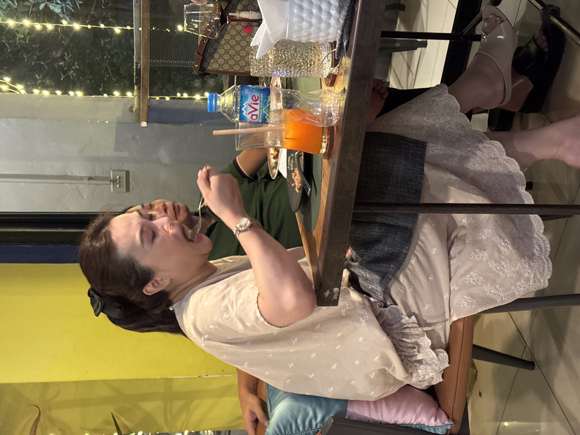 A woman is seated at a table in a restaurant, with her mouth open as if eating, holding a fork in her right hand. She is wearing a light-colored, short-sleeved blouse with a ruffled hem and a patterned skirt. A man in a green shirt is seated to her right. On the table are several drinks, including a bottle of La Vie water, a glass of orange juice, and a glass of water. There are also plates with food, napkins, and a handbag. The restaurant has yellow walls and is decorated with fairy lights. The flooring appears to be tiled. The scene depicts a casual dining experience.