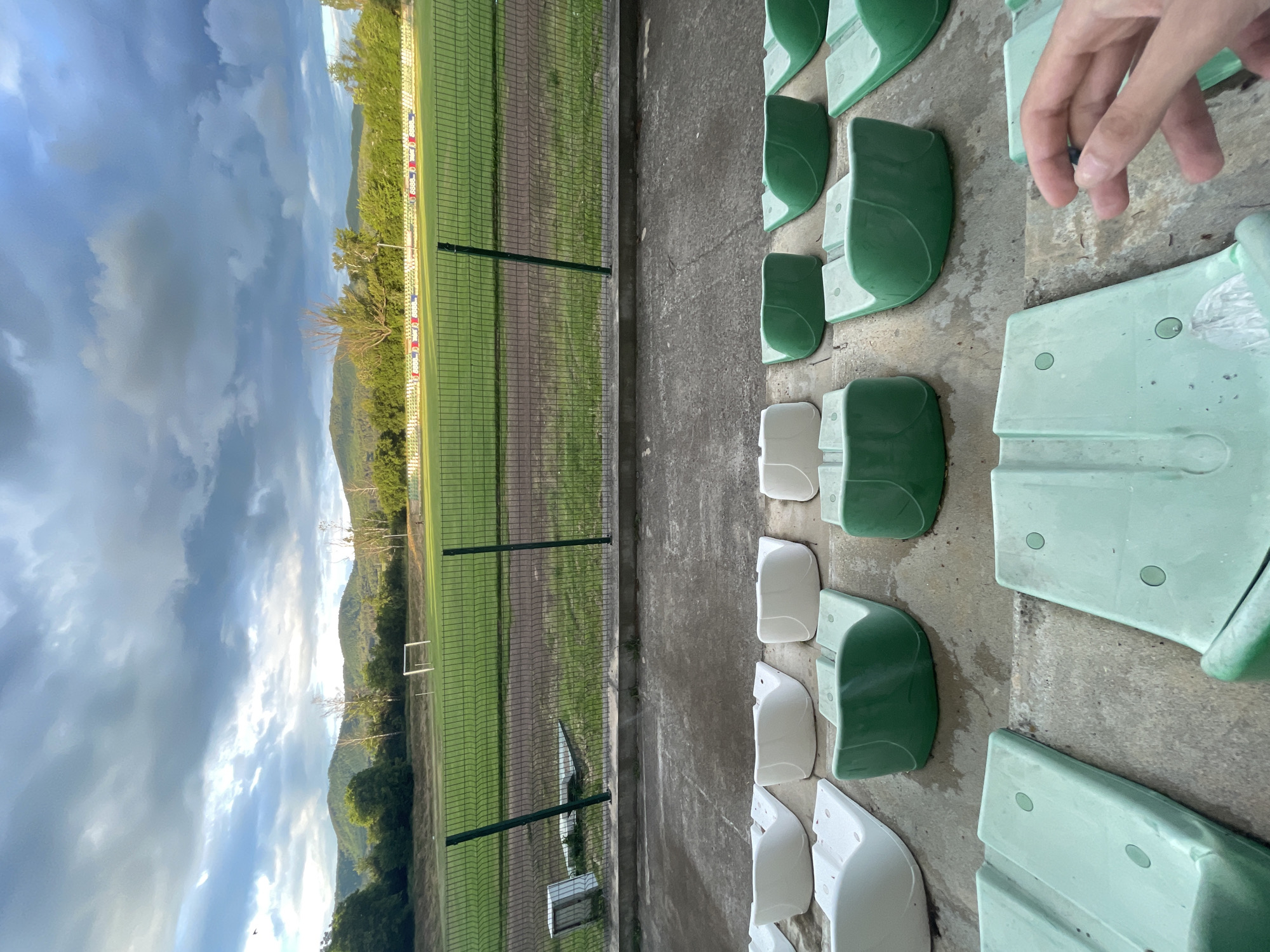 This image captures a football (soccer) pitch and its adjacent spectator seating, viewed from the perspective of someone in the stands. The scene is set outdoors, with a backdrop of green mountains under a partially cloudy sky.
In the foreground, rows of stadium seats are visible, primarily in shades of green, with some white or light gray seats interspersed. These seats appear to be molded plastic, showing some signs of wear. The concrete floor of the seating area is also visible. In the upper right corner, a human hand with light skin is partially shown, holding a small, dark object between its fingers.
The central part of the image features a green grass football field, separated from the seating area by a dark green wire mesh fence. The grass pitch has some visible darker patches, possibly indicating worn areas or recent repairs. A white goal frame is faintly discernible in the distance on the field. Beyond the field and fence, a banner with indistinct text, possibly "OODS" or similar, and small logos is visible, likely indicating sponsors or advertisements.
The background is dominated by a range of verdant, tree-covered mountains and hills that stretch across the horizon. The sky above is largely cloudy but with bright patches, suggesting daytime, possibly late morning or afternoon.
The overall impression is that of a local or smaller sports venue. There is no active game or event taking place on the field. The hand in the foreground suggests a person is present in the stands, observing the empty pitch. The location context is Ihtiman, Bulgaria.