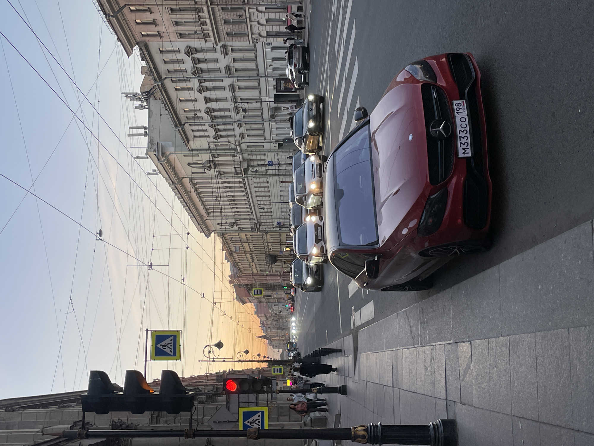 The image captures an urban street scene in Saint Petersburg, Russia, at dusk. The perspective is from a sidewalk, looking down a long, straight avenue lined with ornate, multi-story buildings.
In the immediate foreground, a red Mercedes-Benz car is prominently parked or stopped on the right side of the road, facing away from the viewer. Its license plate, "M333CO 198 RUS," is clearly visible, with "198" indicating the Saint Petersburg region. The sidewalk features large, light-colored paving stones. A black traffic light, showing a red signal, is visible on the left, along with a blue square street sign depicting a yellow arrow pointing upwards with two converging arrows, likely indicating merging lanes.
Further down the street, a queue of vehicles with illuminated headlights is stopped in traffic, stretching into the distance. Several pedestrians are scattered along the sidewalks, some waiting near the traffic light, others walking further away. Overhead, numerous electrical wires crisscross the sky, typical for urban transportation infrastructure. On the rooftops of some buildings, modern cellular antennas are visible, contrasting with the classical architecture.
The sky transitions from a soft blue overhead to warm hues of orange and pink near the horizon, indicating a sunset or the golden hour of the evening. The overall atmosphere is calm, characteristic of a city winding down for the day.