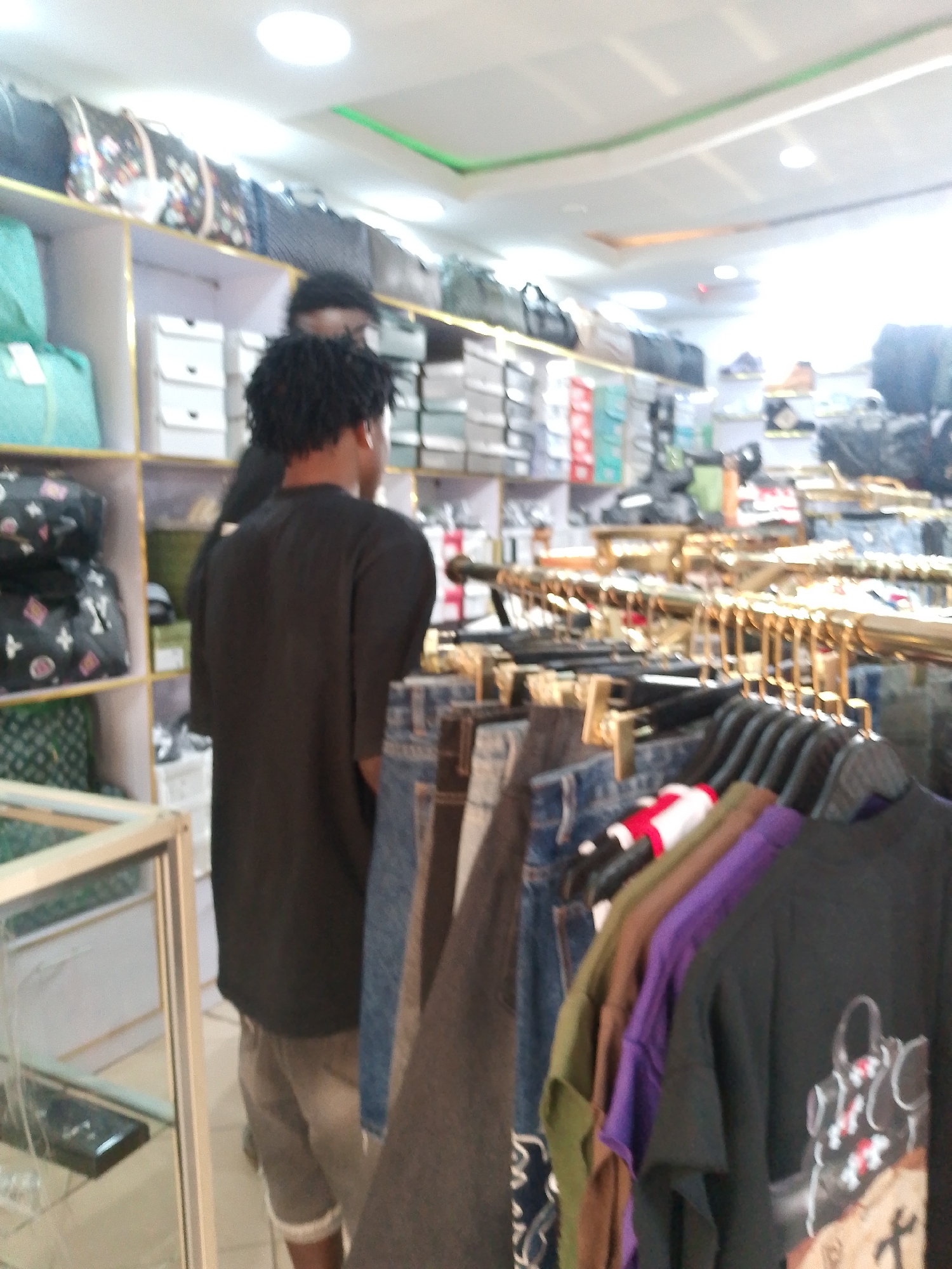 In Bukuru, Nigeria, a retail store displays merchandise on shelves and hanging racks. A person wearing a black t-shirt and grey shorts stands in the foreground, facing away from the camera. Another person is partially visible behind them. The store environment features various items including bags on shelves, folded boxes, and a display cabinet. A rack of colorful t-shirts hangs in front of the person, suggesting an apparel or fashion retail setting.