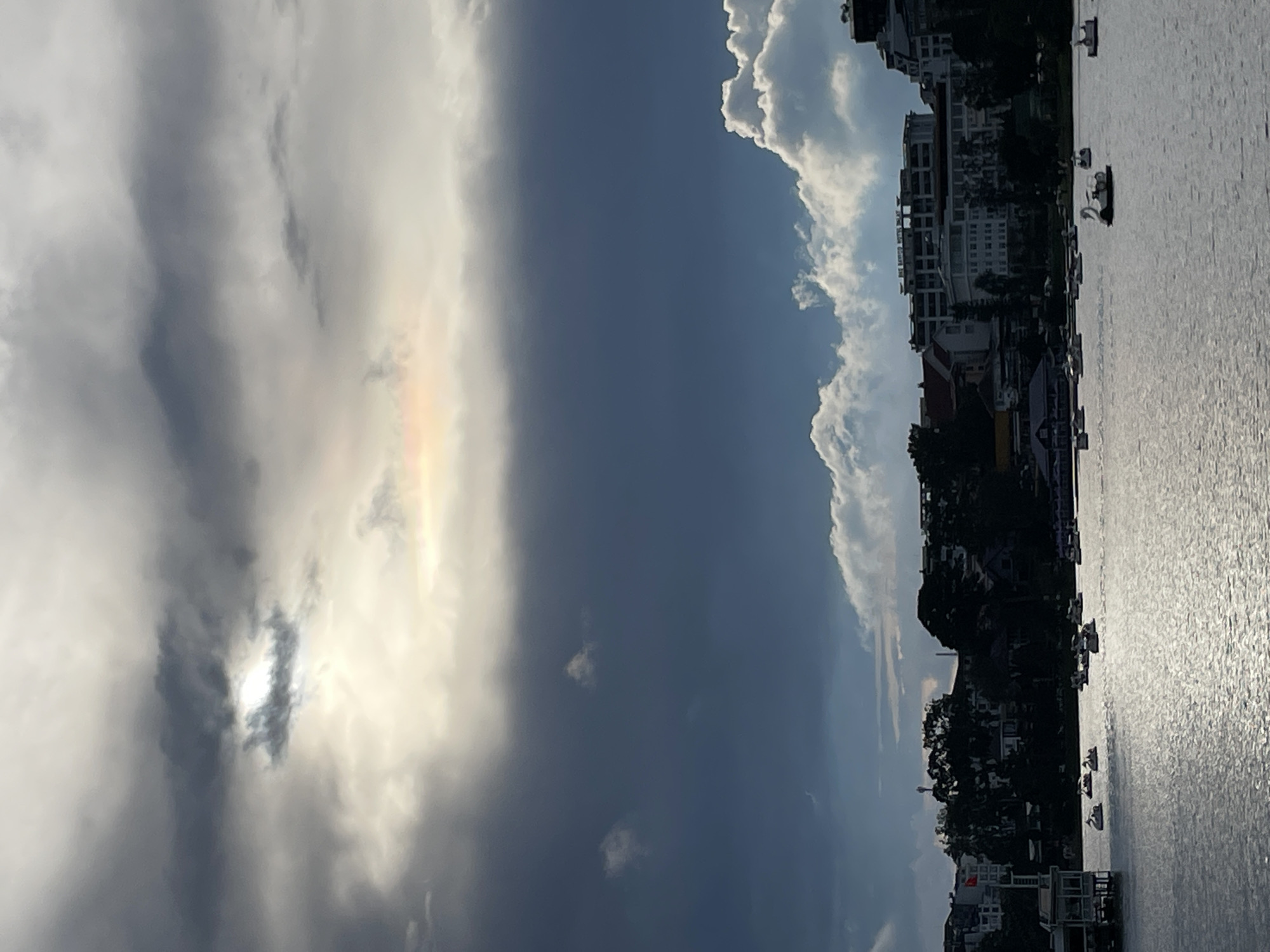 The image depicts a wide shot of Xuan Huong Lake in Da Lat, Vietnam, under a dramatic sky. The water is calm and reflects the light, with numerous swan-shaped pedal boats scattered across its surface. In the foreground, the lake occupies the majority of the frame. The middle ground features a shoreline lined with trees and buildings, including what appears to be the Dalat Palace Hotel. The background is dominated by a cloudy sky, with a break in the clouds allowing sunlight to illuminate a portion of the sky with a faint rainbow. The overall mood is serene, with a hint of impending weather due to the dark clouds.