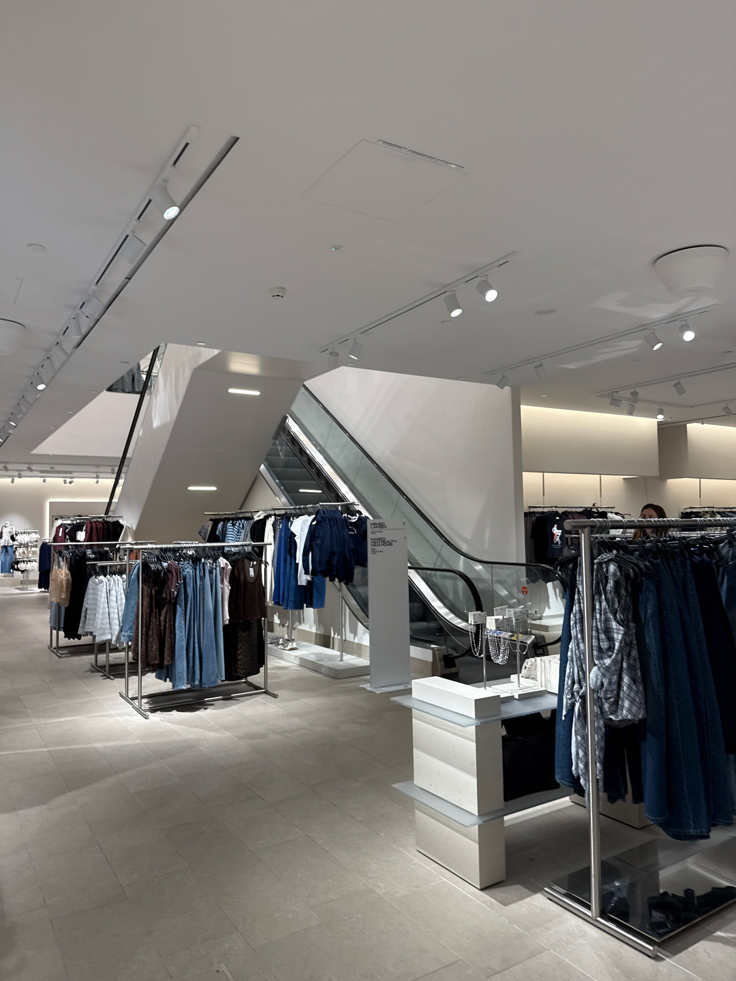 This is a photograph taken inside a retail store, likely a clothing store, with a modern and minimalist design. The store appears to be well-lit with recessed ceiling lights and track lighting. The floor is tiled with large, light-colored tiles.
In the center of the image, a prominent staircase and escalator ascend to an upper level. The architecture of the staircase is sleek and angular, made of white material. Clothing racks are visible throughout the store, displaying a variety of garments. Some racks are filled with dresses and skirts in denim and darker fabrics, while others feature blouses and jackets. The clothing appears to be neatly arranged.
In the foreground, to the right, is a display unit with stacked elements and glass shelves, showcasing jewelry and small accessories. A person, partially visible, is standing behind a clothing rack on the right side of the image, looking towards the left. Their face is partially obscured, but they appear to be a young woman with brown hair.
The overall impression is that of a contemporary retail environment, likely a mid-range to high-end fashion store. While the specific location within Tallinn, Estonia, cannot be determined from the image alone, the scene depicts a typical commercial interior found in a modern city.