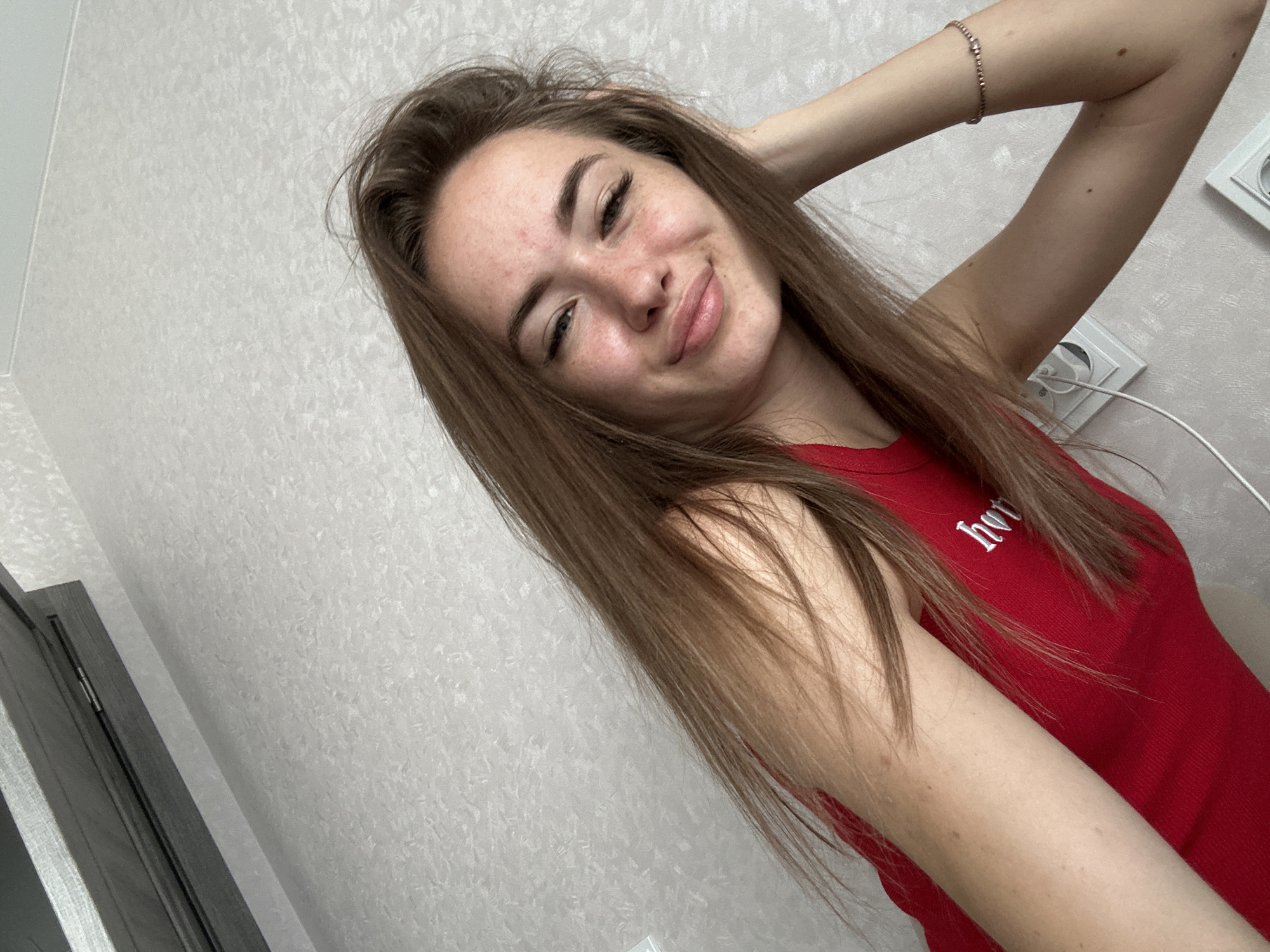 A young woman with long brown hair and freckles poses for a selfie in a room with textured wallpaper. She is wearing a red top with white lettering. Her right arm is raised, and she is touching her head. A delicate bracelet adorns her left wrist. Two electrical outlets are visible on the wall behind her.