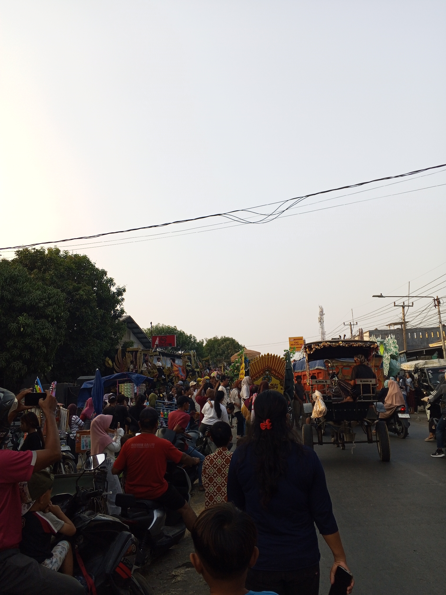 The image depicts a street scene in Gebang, Indonesia, possibly during a parade or festival. A horse-drawn cart, adorned with decorative elements, is the central focus of the scene, moving down a paved road. Several people are visible, some on motorcycles, others standing and observing. Trees line the left side of the street, and various stalls or temporary structures are set up, suggesting a marketplace or event. The sky is overcast with a pale, hazy light. The presence of numerous people indicates a public gathering.