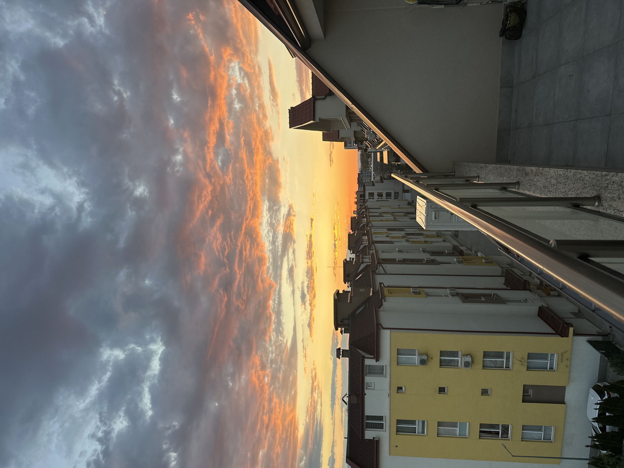 The image shows a view from a balcony overlooking a row of apartment buildings in Plovdiv, Bulgaria. The buildings are mostly white with some yellow sections and red-tiled roofs. Air conditioning units are visible on the buildings. A metal railing and a portion of the balcony's concrete floor are in the foreground. The sky is filled with clouds, displaying a sunset with colors ranging from grey to orange. A glimpse of a car and some greenery are seen at the bottom of the image.