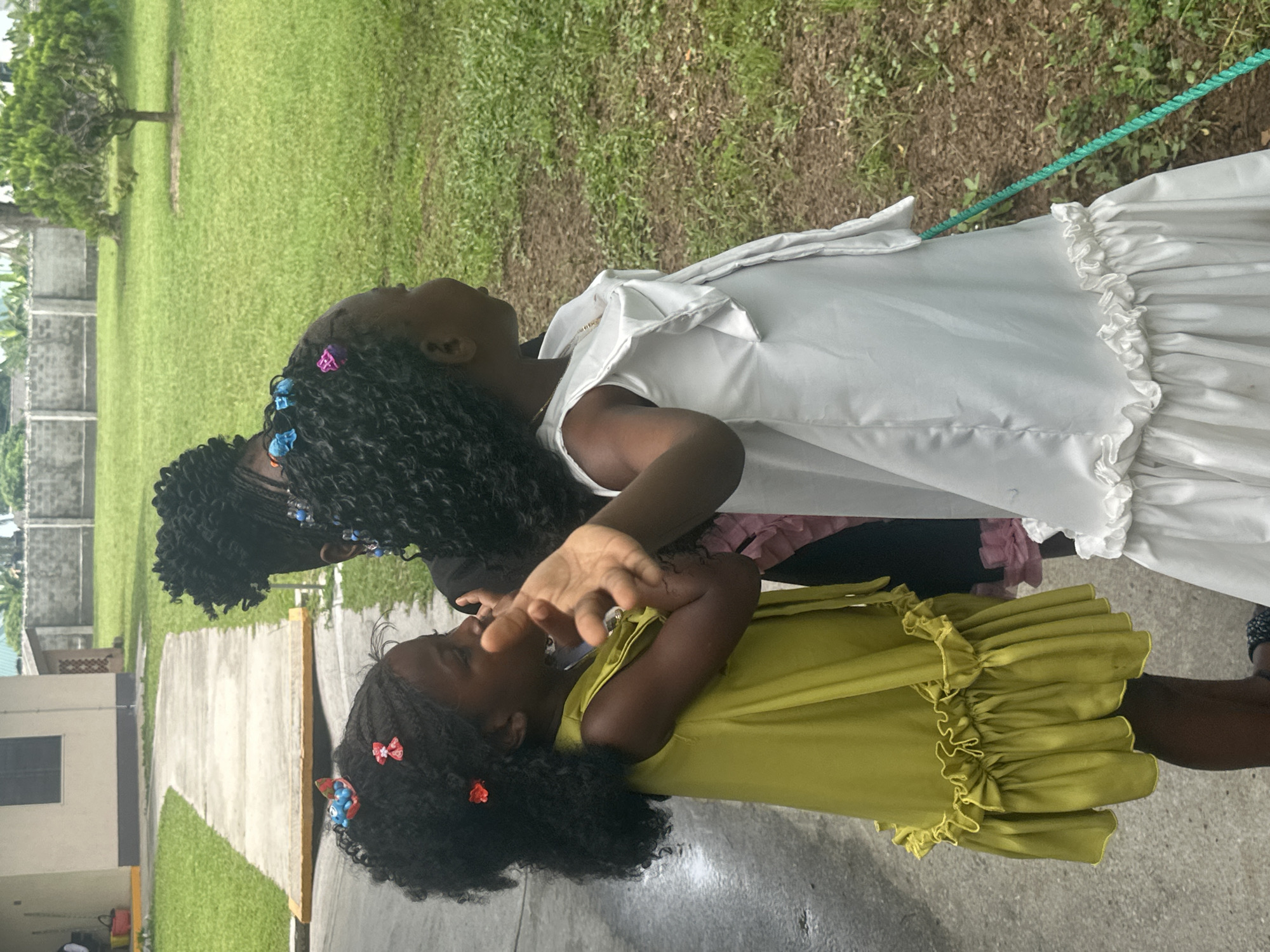 The image shows three young Black girls standing outside, with two of them in the foreground and one partially visible behind them. The girl on the left is wearing a bright yellow dress with ruffled trim at the hem and has her hair styled in braids adorned with colorful hair clips. She is looking to her left and her left hand is raised, appearing to interact with something just out of frame or to be posing. The girl in the center, wearing a white dress with a ruffled hem and a necklace, is looking to her right, with her arm resting on the shoulder of the girl in yellow. The third girl, visible behind the other two, has her hair in braids as well and is facing away from the camera. They are standing on a paved surface next to a grassy area. In the background, there is a light-colored building with a dark window, a concrete wall, lush green grass, and a small tree. The overall setting suggests an outdoor environment, possibly a school or residential compound in Itoga, Nigeria.